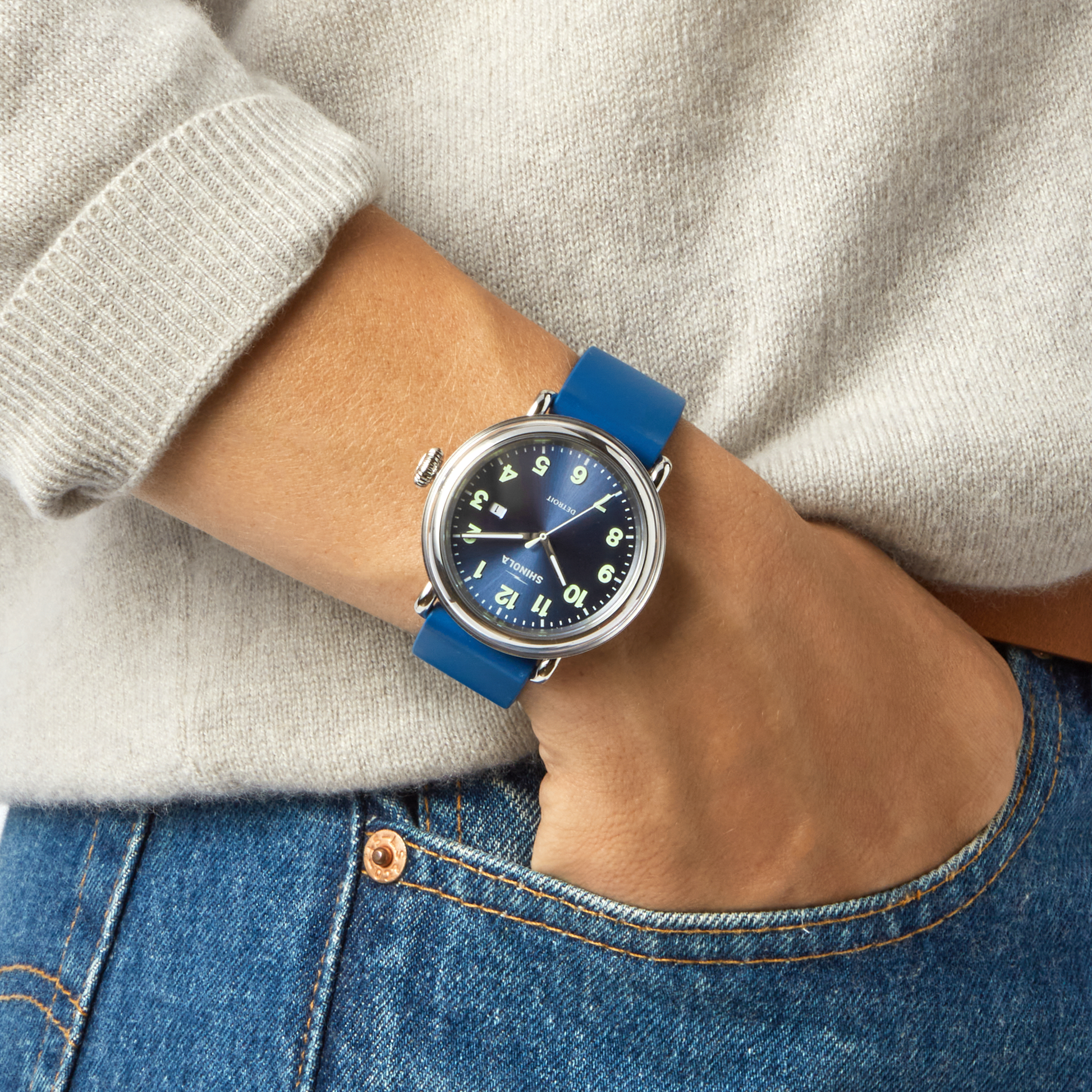 A person wearing the Shinola Detrola Watch - Denim Blue, featuring a round face with white numbers and a blue strap, styled with a light sweater and blue jeans, one hand casually in their pocket.