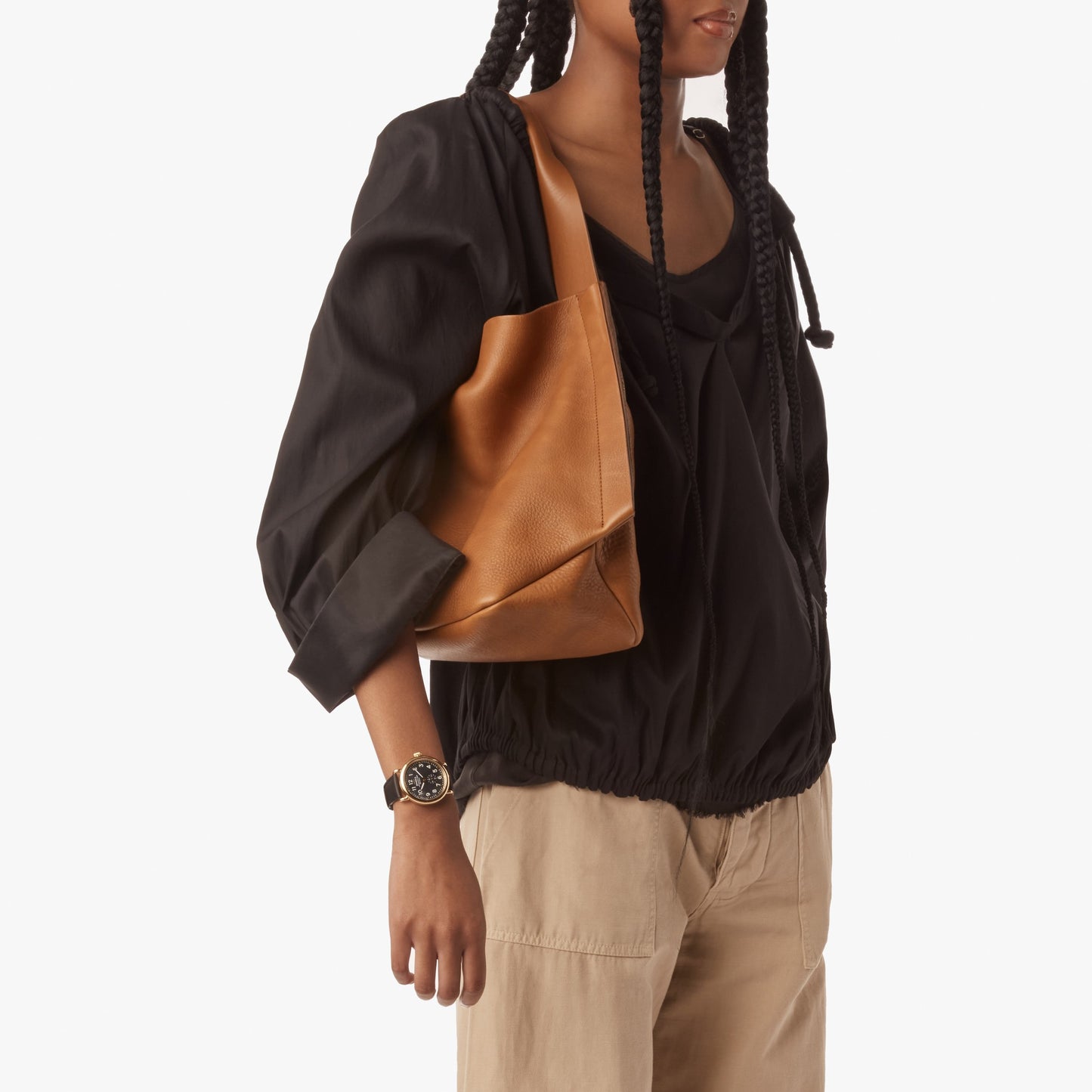 A person with long braids wears a loose black top, beige pants, a watch, and carries the Shinola Derby Shoulder Bag - Tan with leather card pocket against a plain white background.