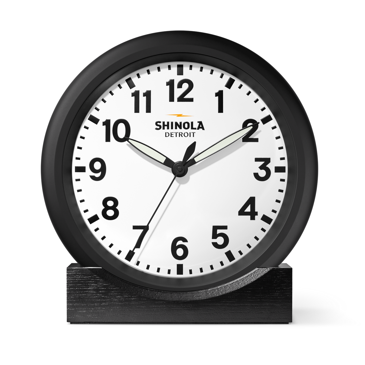 The Shinola 14" Runwell Clock - White features a round white face with black numbers and hands, a black rim, and displays 1:51:33. It sits on a black stand with "Shinola Detroit" on its face.