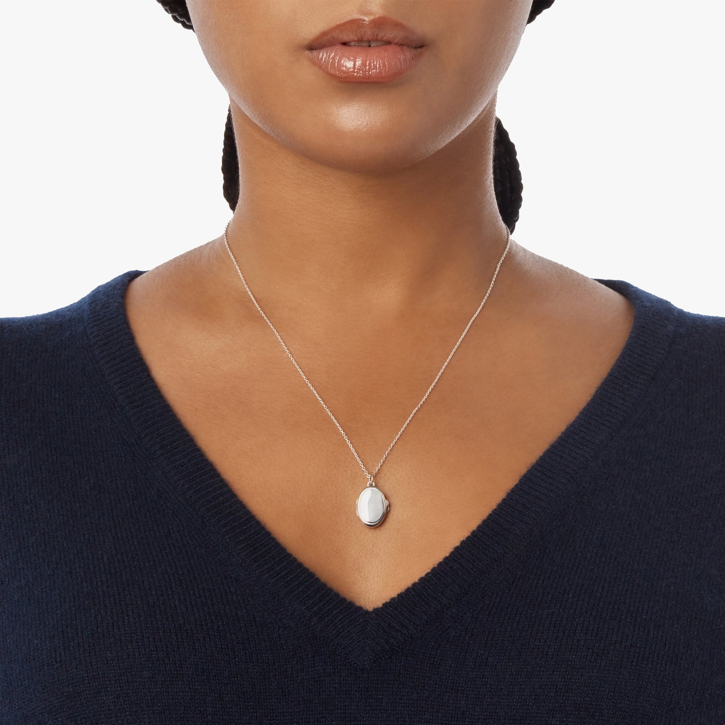 Shown from shoulders to lips against a white background, a person wears a navy V-neck sweater and the Shinola Heirloom Petite Locket Necklace in sterling silver.