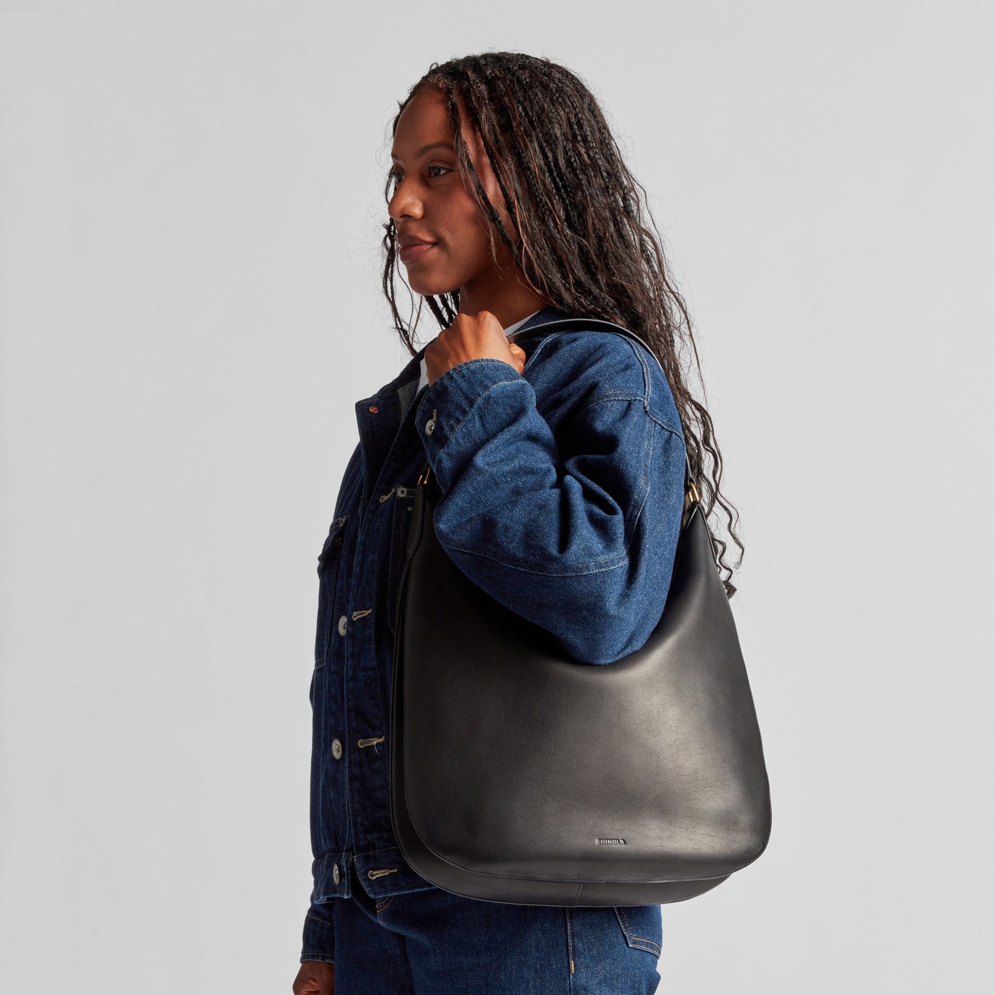 A woman in a blue denim jacket stands in profile, holding the Shinola Bixby Shoulder Bag - Black with an accordion-style design, against a plain gray background.