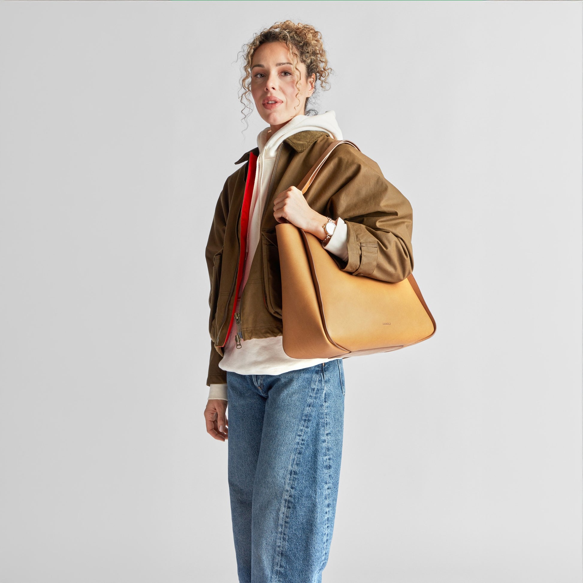 A woman with curly hair, dressed in a brown jacket, white hoodie, and blue jeans, stands with the Shinola Derby Tote Bag - Tan made from natural leather over her shoulder against a plain light gray background.
