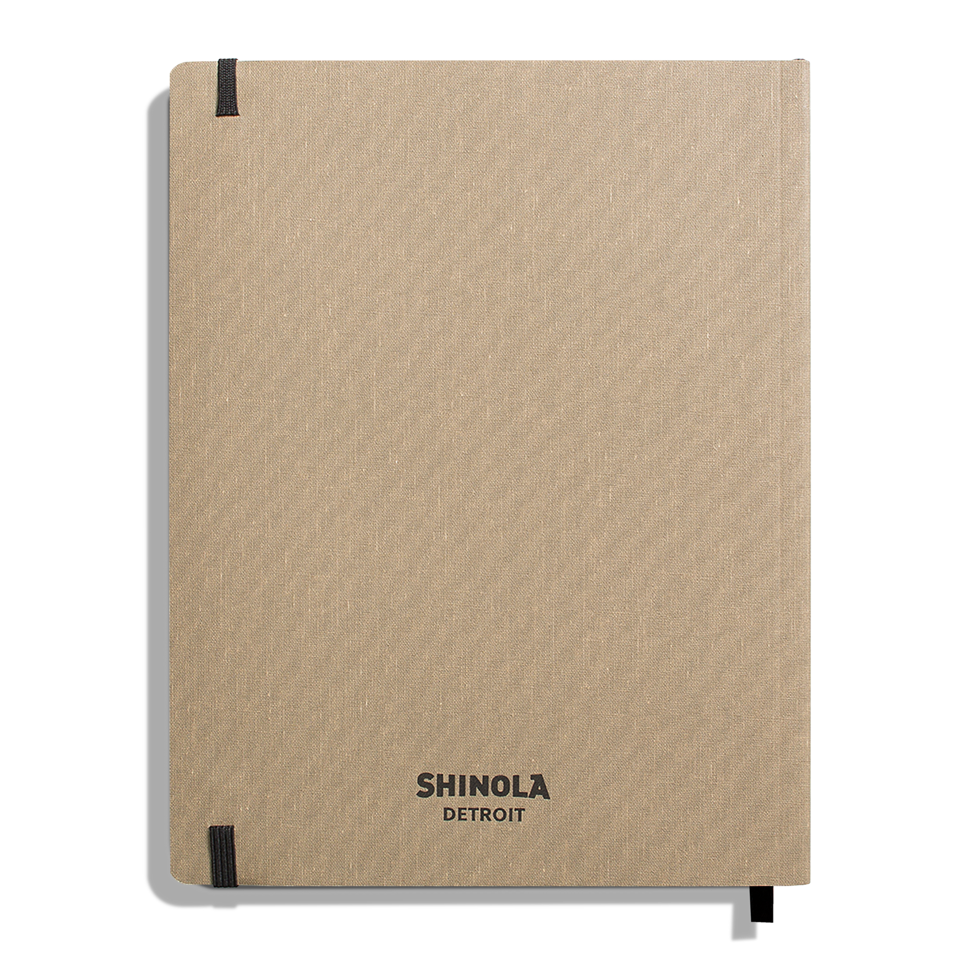 A large, oat-colored softcover ruled journal by Shinola, American-made, with black elastic bands at two corners and “Shinola Detroit” in black at the bottom center of the cover.