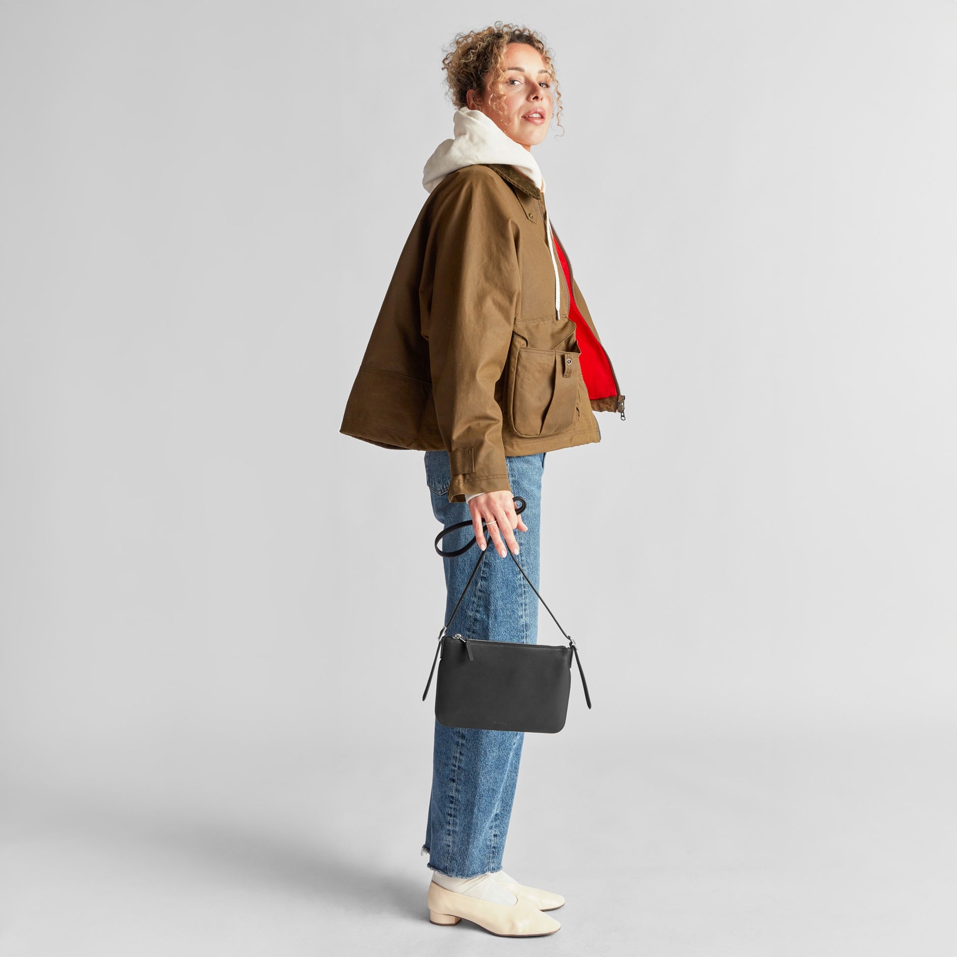 A person in profile wears a brown jacket, blue jeans, white boots, and a white hoodie, holding the Shinola Small Bixby Crossbody - Black and looking toward the camera against a plain background.