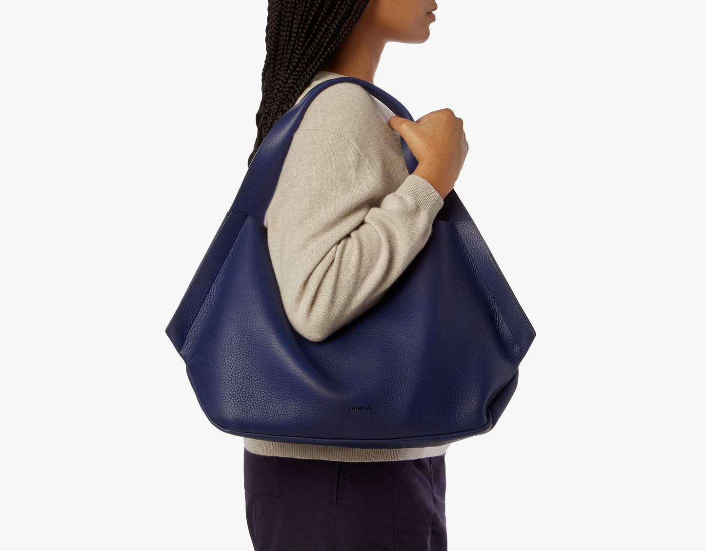 A person with braided hair, in a beige sweater and dark pants, carries the Shinola Runwell Shoulder Bag - Lake with carrying handles and a leather card pocket on their right shoulder against a plain white background.
