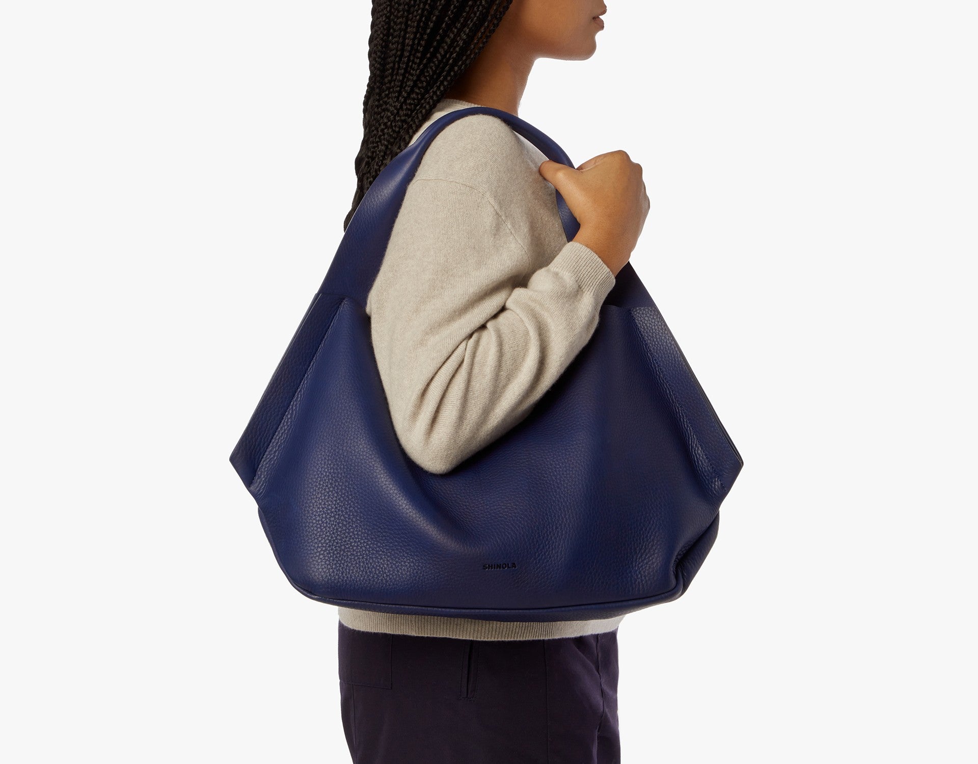 A person with braided hair, in a beige sweater and dark pants, carries the Shinola Runwell Shoulder Bag - Lake with carrying handles and a leather card pocket on their right shoulder against a plain white background.