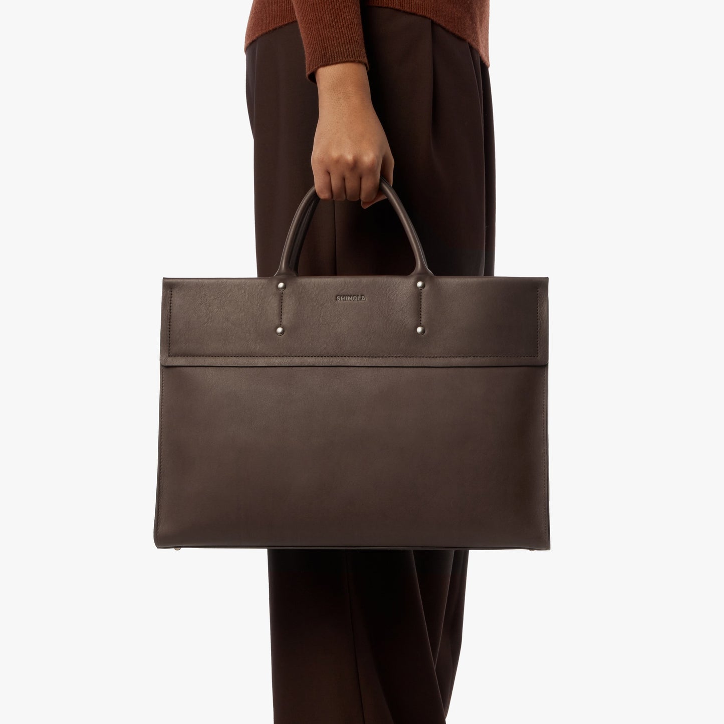 A person holds the Shinola Day Satchel - Espresso by its handles against a plain white background, wearing brown pants and a brown top.