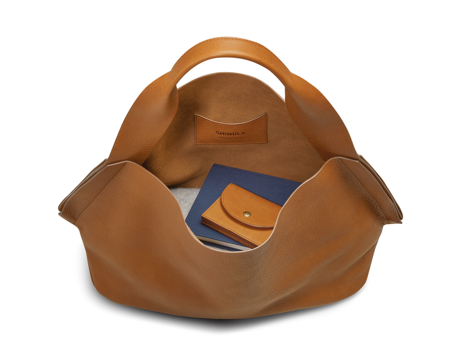 The Shinola Derby Shoulder Bag in tan, shown open with carrying handles, reveals a blue notebook, tan leather wallet, and glasses inside. Its interior includes a leather card pocket stamped “Shinola.”.