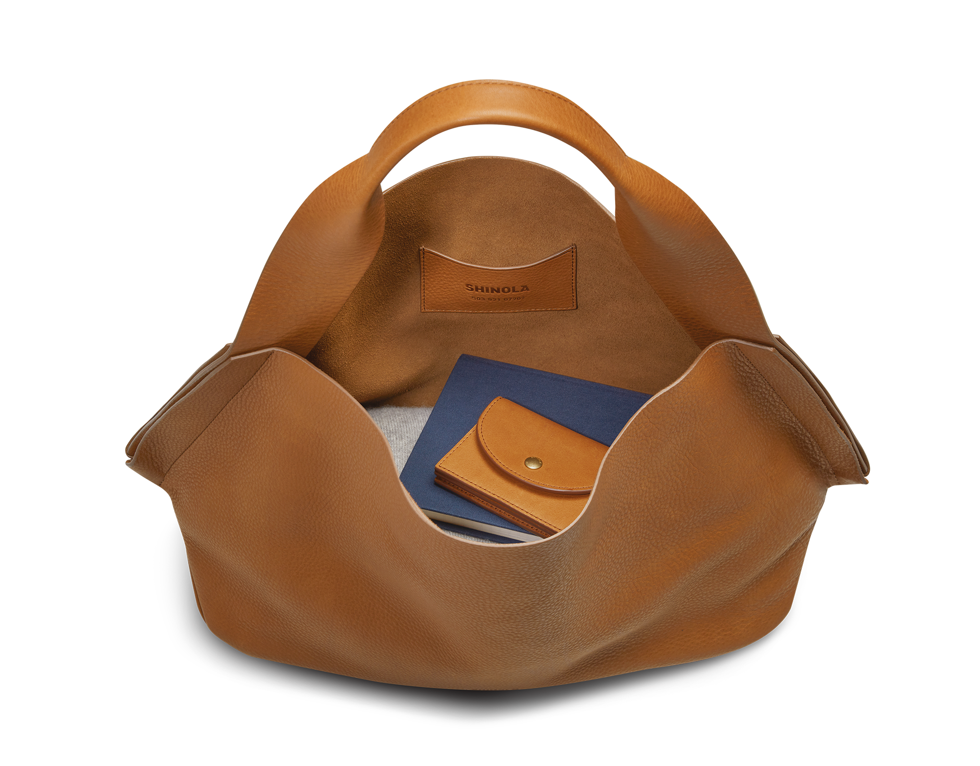The Derby Shoulder Bag - Tan by Shinola is open, displaying a blue notebook, tan leather snap-button wallet, and glasses. The interior pocket serves as a leather card pocket embossed with the Shinola brand name.