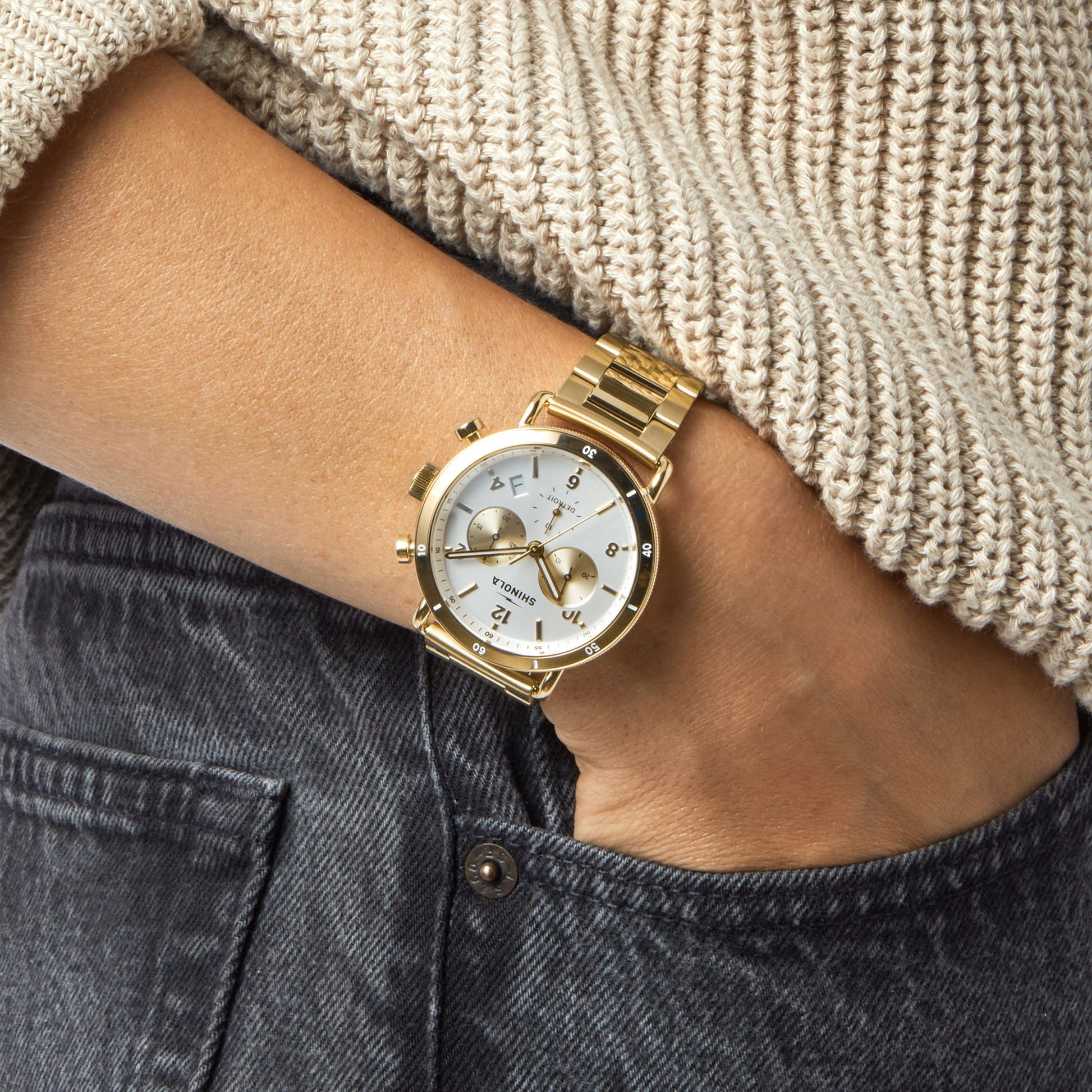 Wearing a knit beige sweater and dark jeans, a person has a Shinola Canfield Sport Chronograph Watch - Silver on their left wrist, its white face and multiple dials visible as they tuck their hand into their jeans pocket.