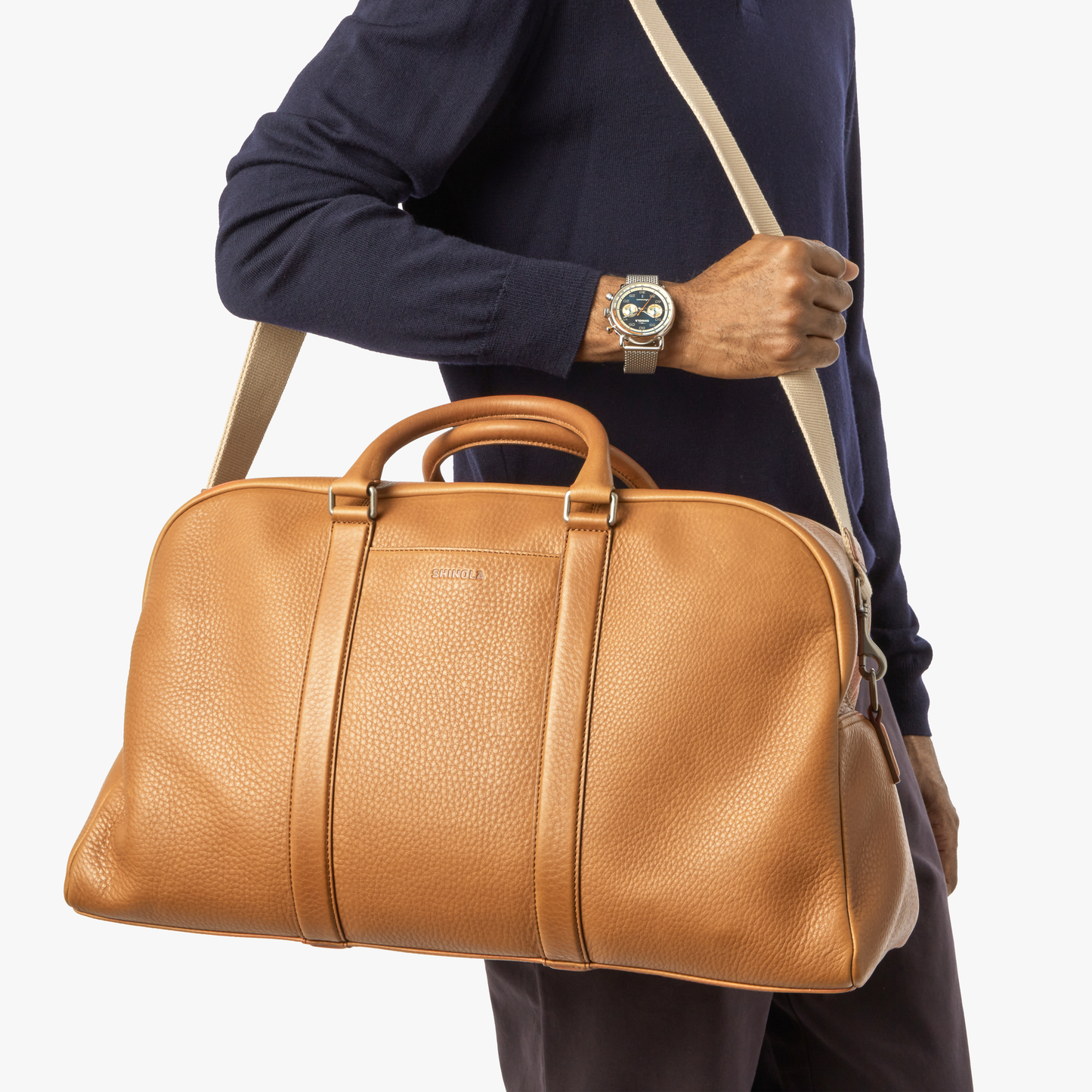 A person in a navy long-sleeve shirt holds the Shinola Runwell Duffle Bag - Tan, featuring a beige shoulder strap, ideal as a TSA carry-on. They wear a wristwatch on their left hand against a white background.