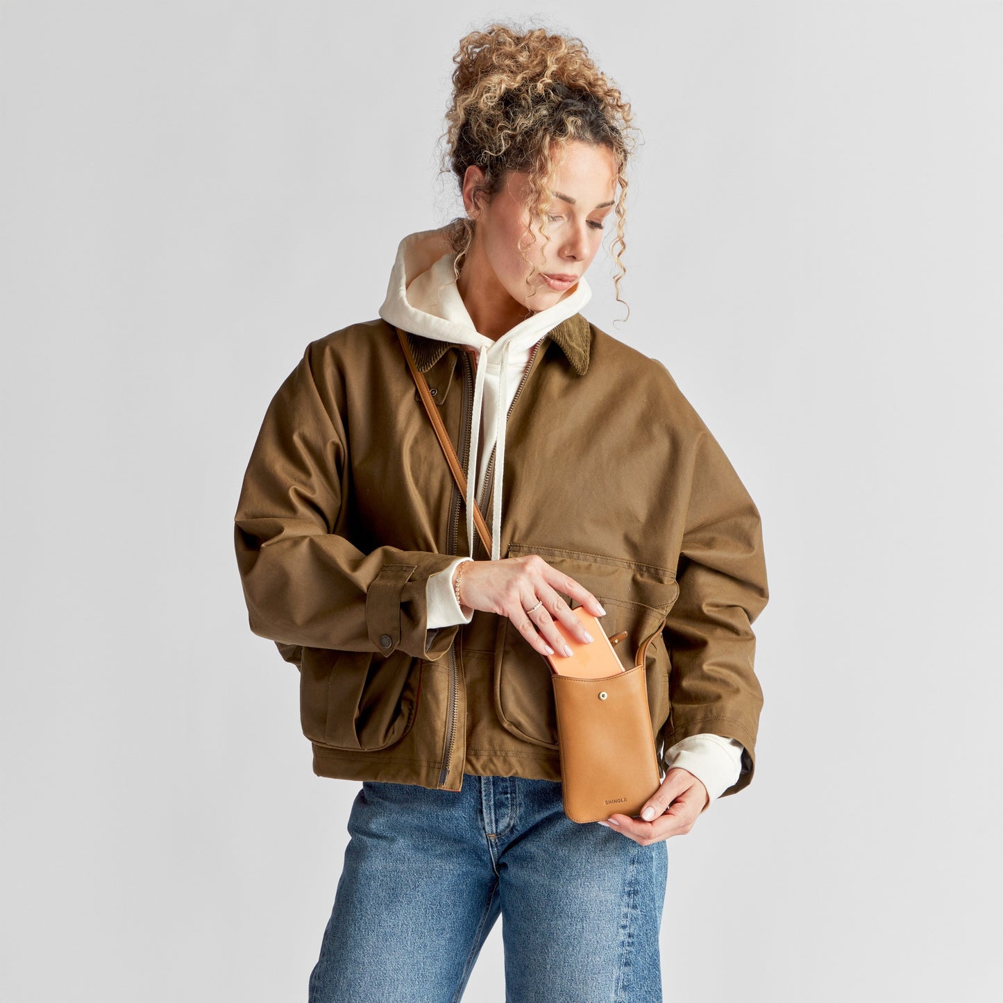 A person with curly hair in a brown jacket, white hoodie, and blue jeans stands against a plain background, placing their hand into the Bixby Phone Crossbody - Tan by Shinola—ideal for traveling light.