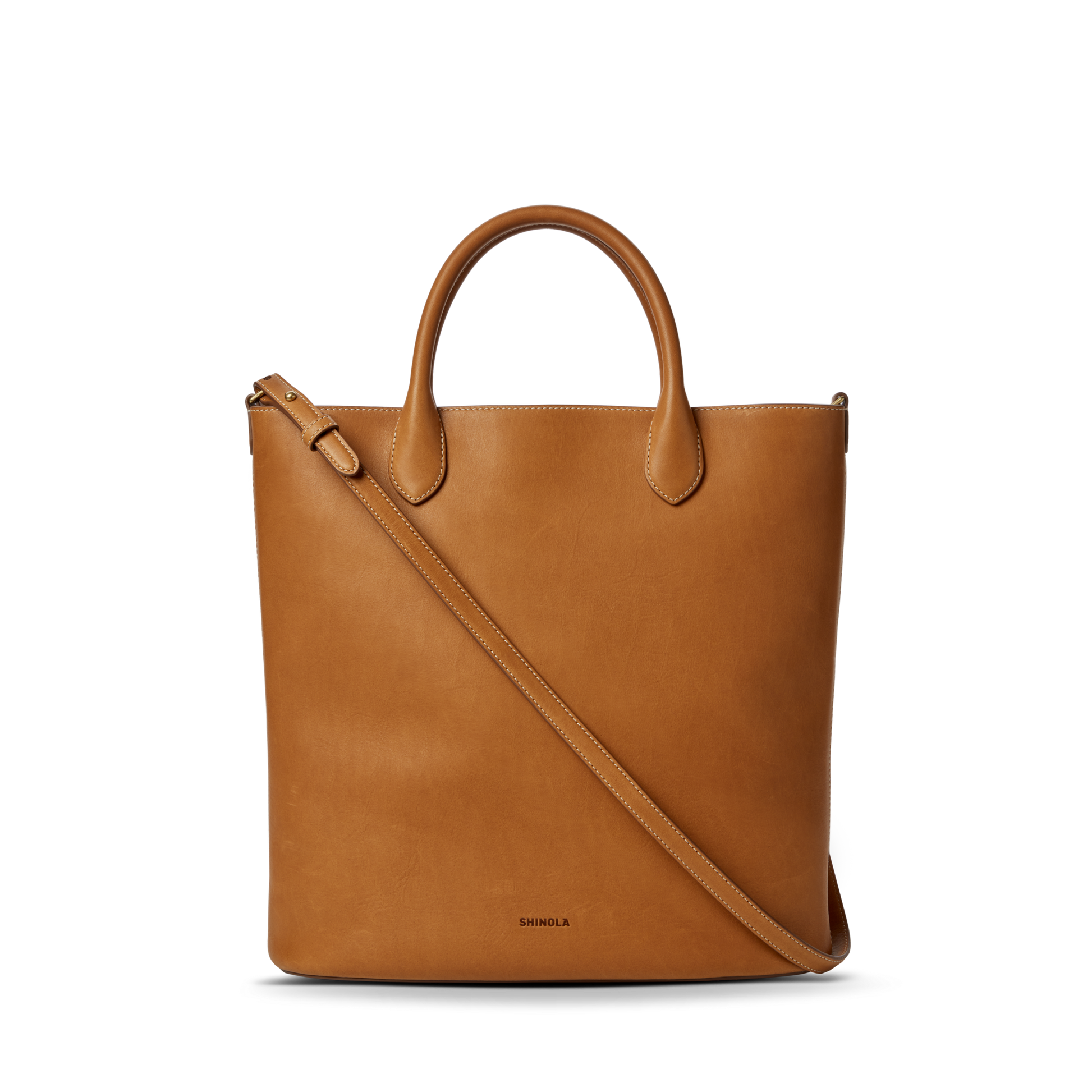 The Shinola 12 Hour Tote Bag in tan features two top handles, a long adjustable shoulder strap, and an interior pocket for organization. "Shinola" is embossed near the bottom center and it's displayed on a plain white background.