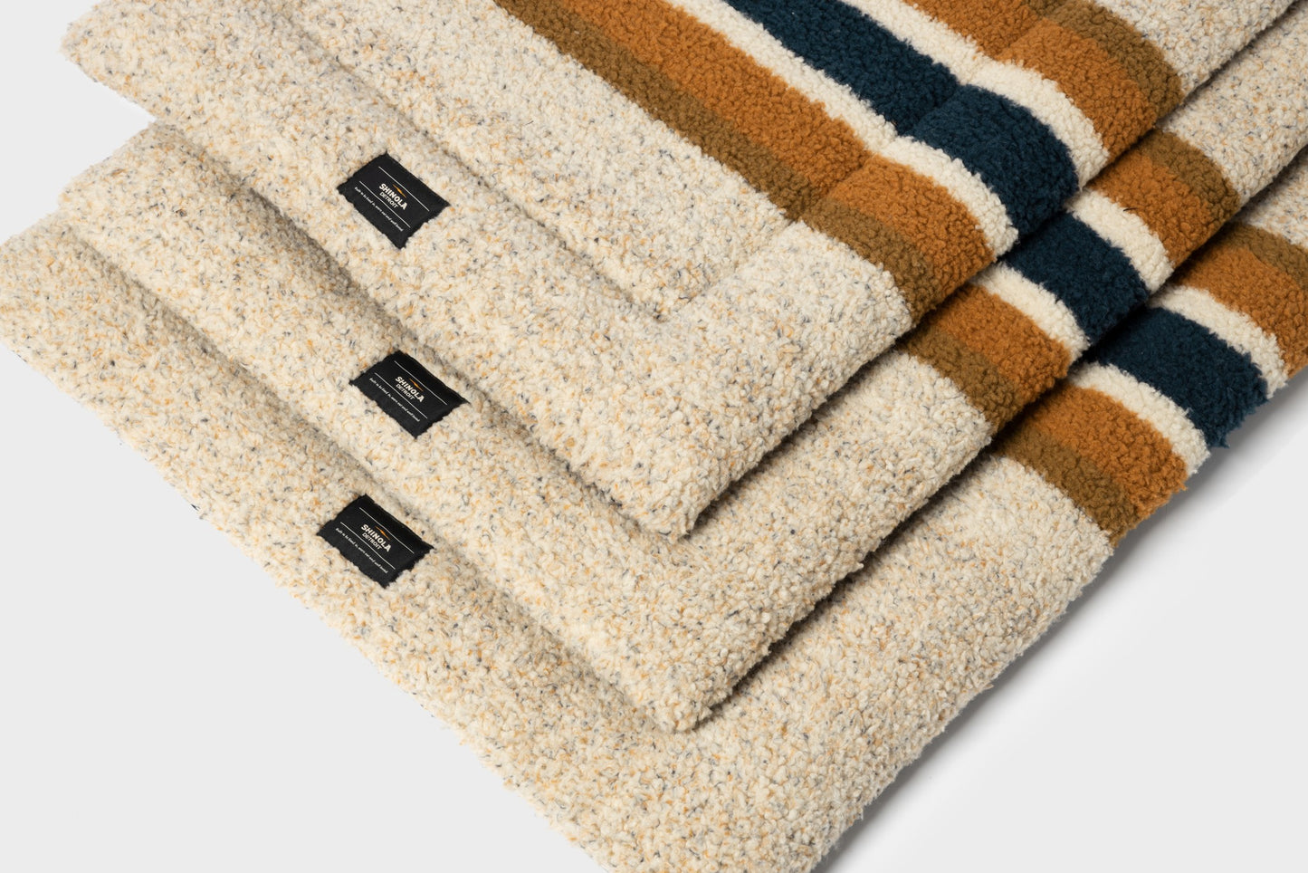 Three folded Shinola Crate Comfort Mat - Oat Stripe wool blankets with cream, brown, and dark green stripes, each with a black corner label—ideal as cozy, machine-washable dog beds or crate cushions—arranged on a light surface.