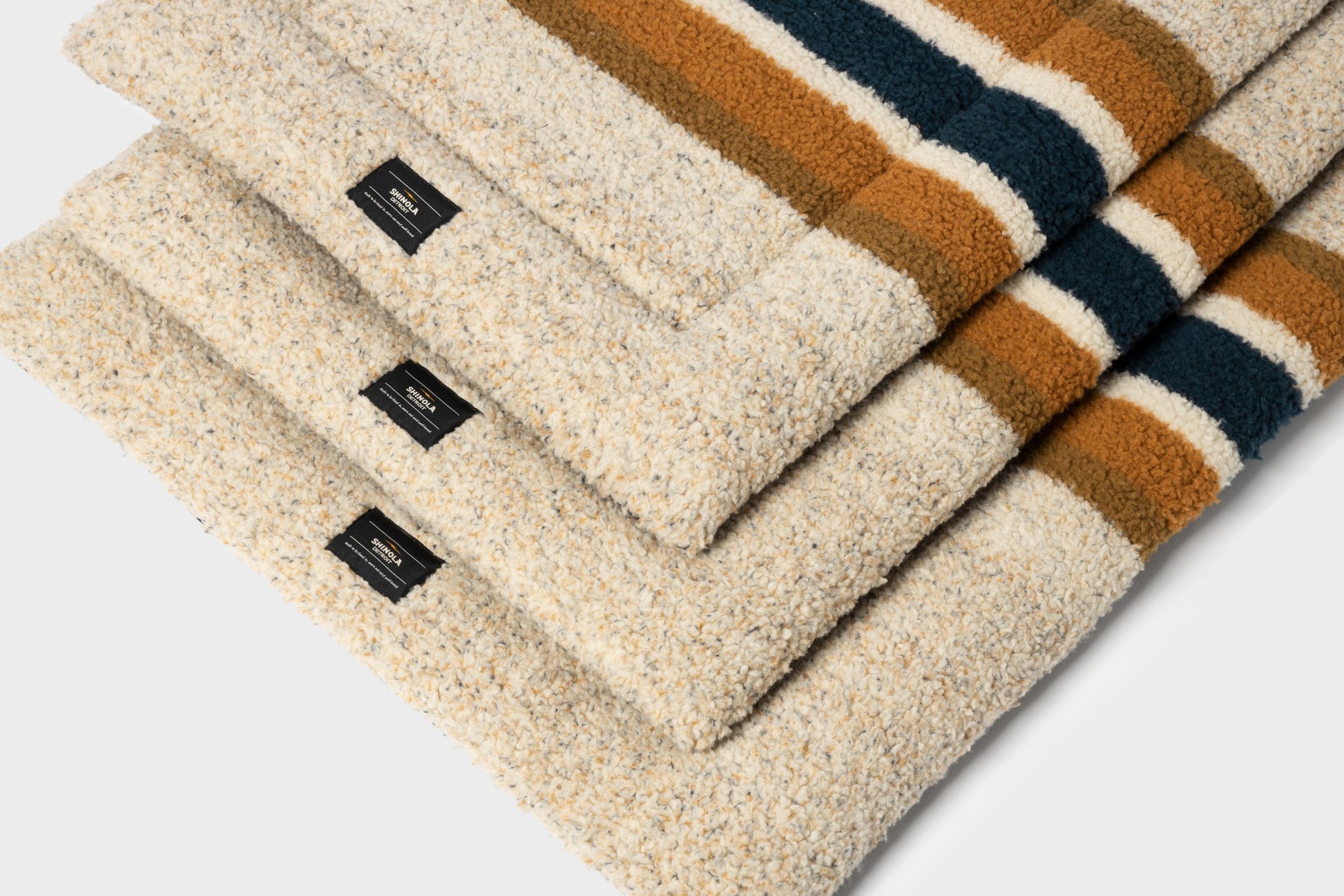 Three folded Shinola Crate Comfort Mat - Oat Stripe wool blankets with cream, brown, and dark green stripes, each with a black corner label—ideal as cozy, machine-washable dog beds or crate cushions—arranged on a light surface.