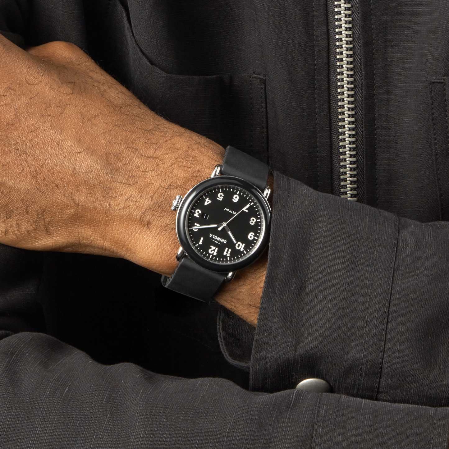 A person wears a black jacket and a Shinola Detrola Watch - Black—featuring a round face, white numerals, and black band—on their left wrist, with their right hand resting on their left forearm.