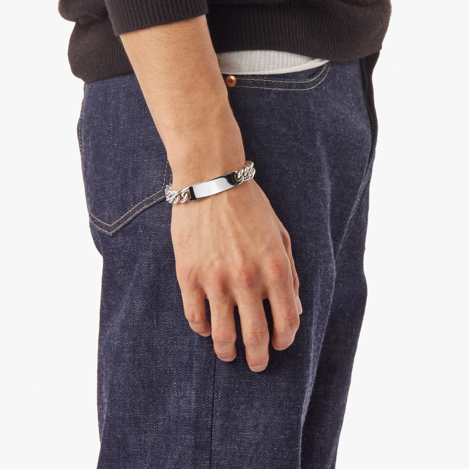 A person in blue jeans and a black sweater, shown from the waist down with their left hand by their side, wears the Shinola Bike Lock ID Bracelet in sterling silver, featuring an engravable blank rectangular plate.
