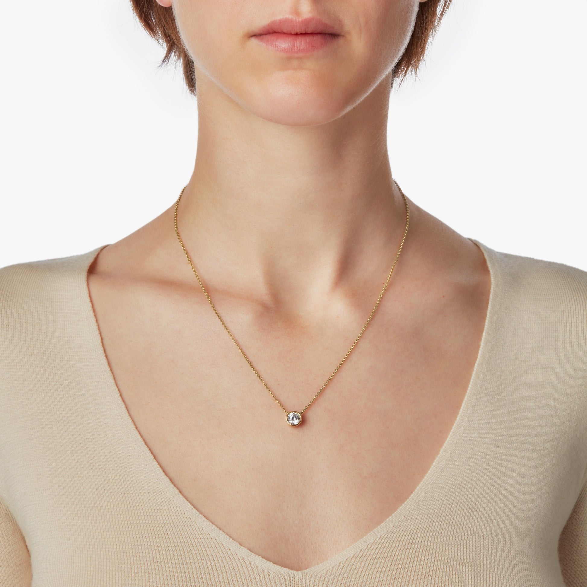 A woman in a cream V-neck top showcases the Shinola 1 Carat Round Diamond Necklace in yellow gold, featuring an ethically sourced bezel-set diamond pendant, centered on her chest against a plain white background.
