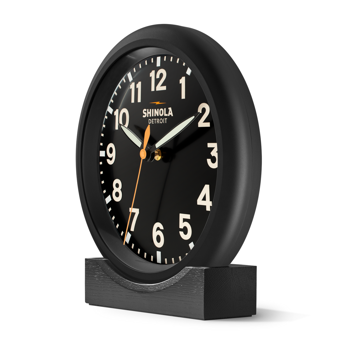 The Shinola 14" Runwell Clock features a matte black dial with white numerals and an orange second hand, elegantly displayed on a black wooden base.