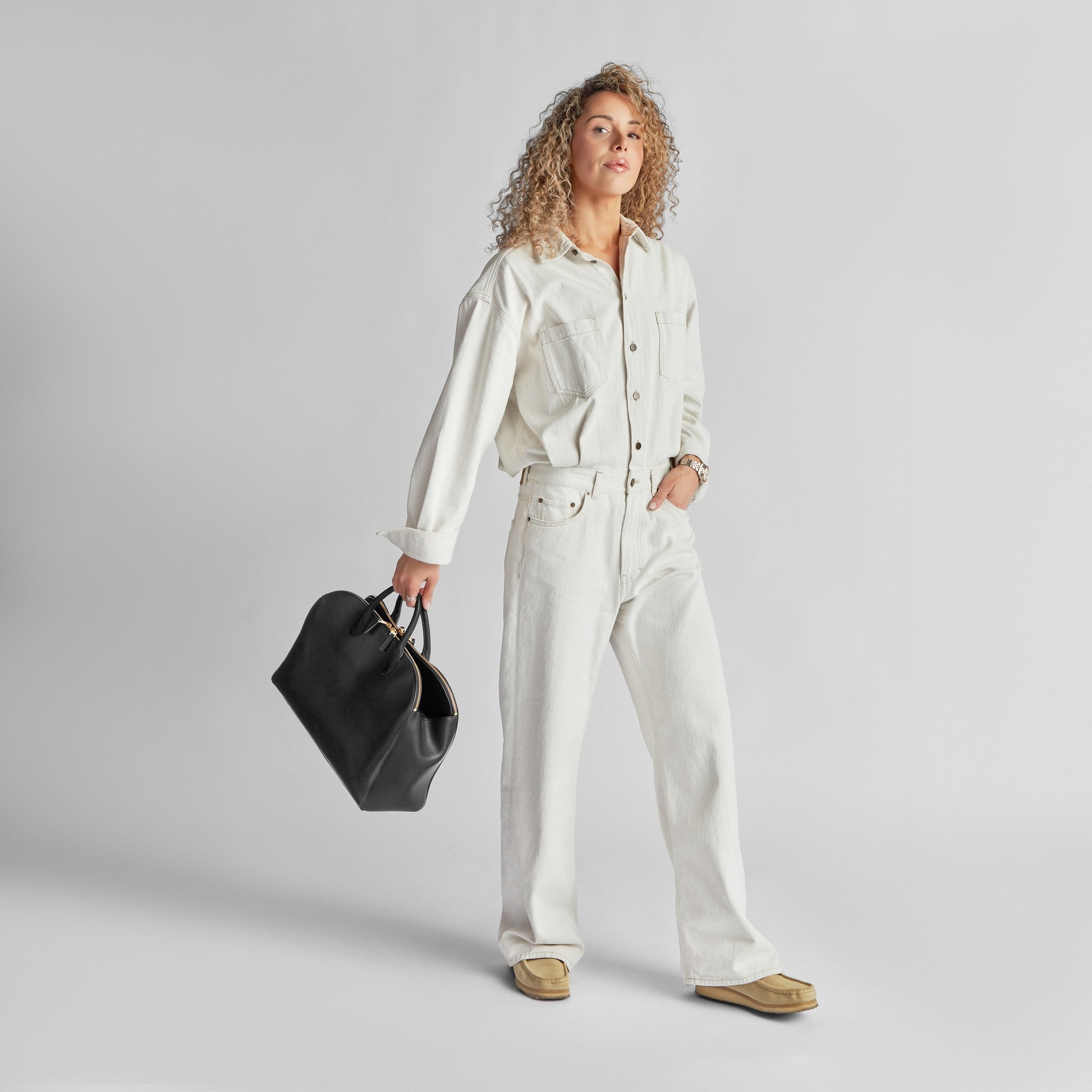 A confident person with curly hair, dressed in white, holds a Shinola Bixby Work Satchel - Black—crafted from natural leather—against a plain light background.