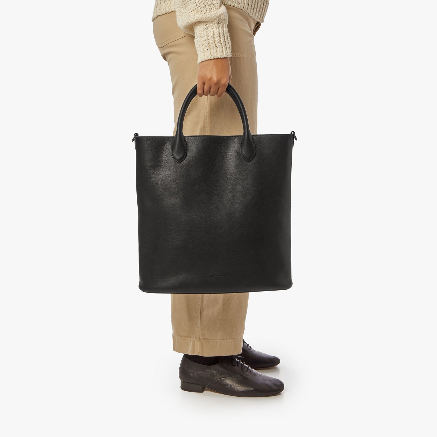 A person in beige pants, a cream sweater, and dark shoes stands against a white background holding the Shinola 12 Hour Tote Bag - Black, a large leather tote designed for everyday use, by its handles.