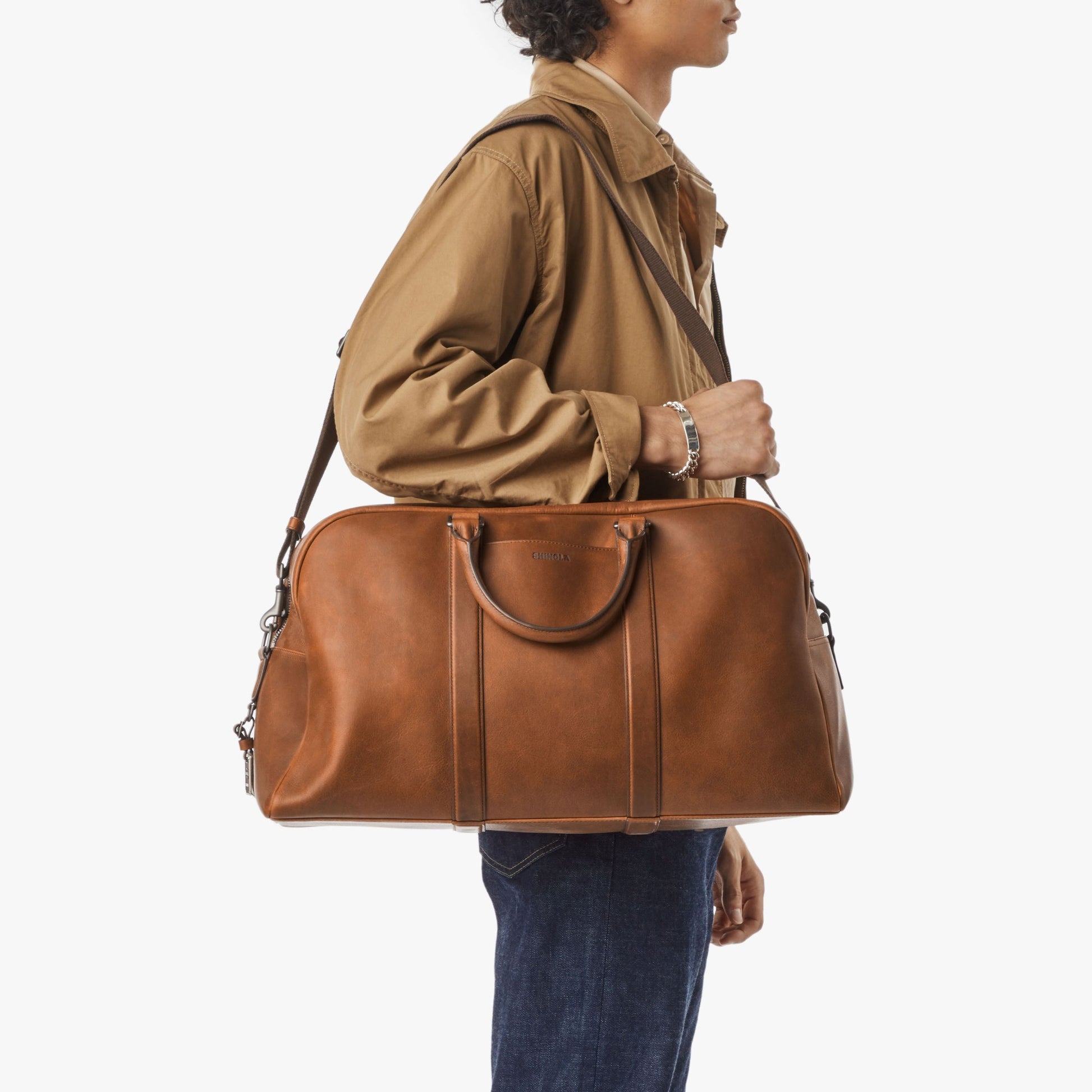 A person in a tan jacket and blue jeans carries the Shinola Runwell Duffle Bag - Medium Brown over their shoulder against a plain white background.