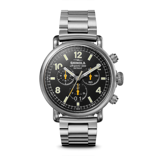 The Shinola Runwell Chronograph West Wind Watch Gift Set features a stainless steel case, black dial with silver hands and yellow accents, three subdials, a metal link bracelet, water resistance to 20 ATM, plus an extra strap. Shown on white.