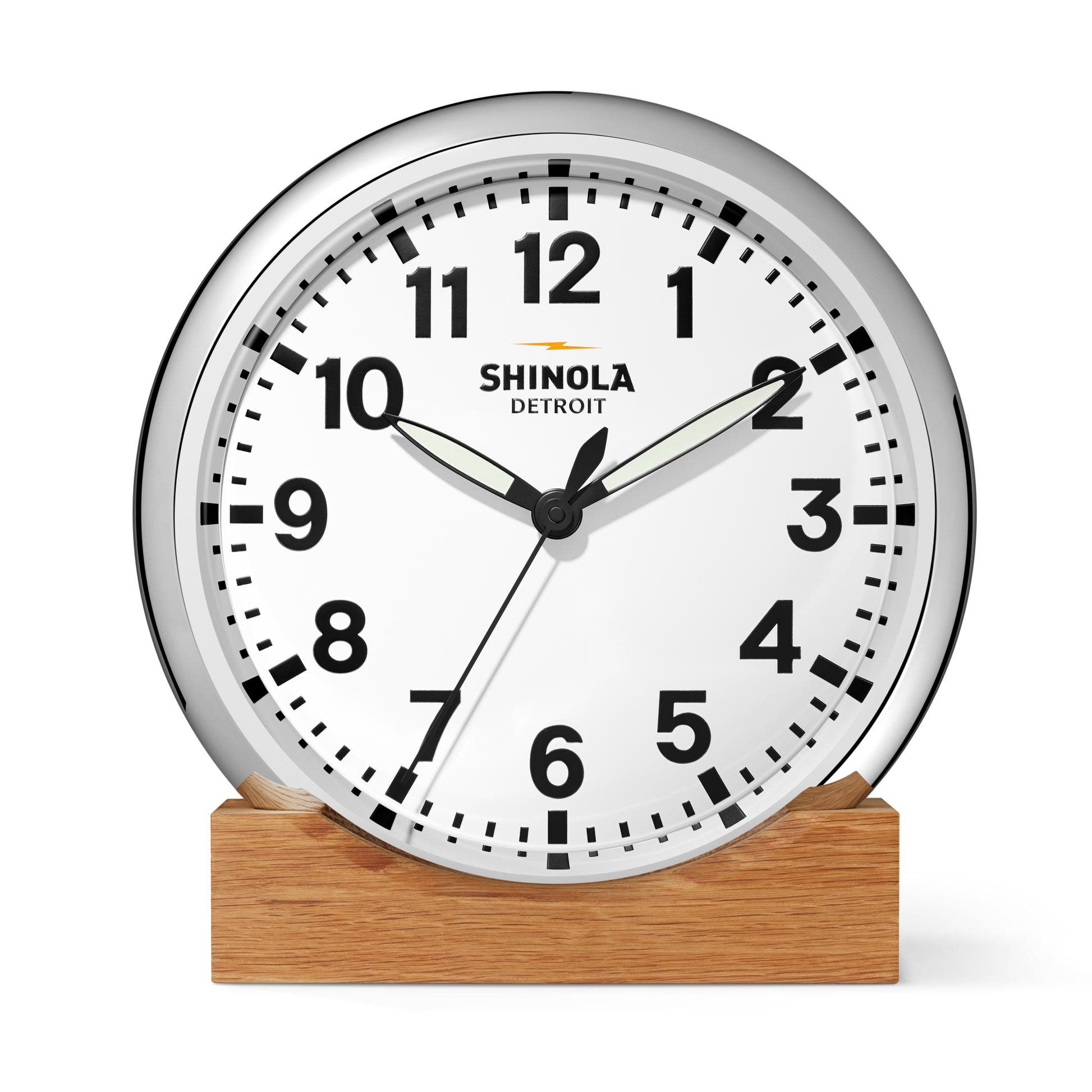 The Shinola 14" Runwell Clock features a white face, black hands and numbers, a chrome casing on a wooden base, and "Shinola Detroit" printed on the dial. It displays the time as 1:50:34.
