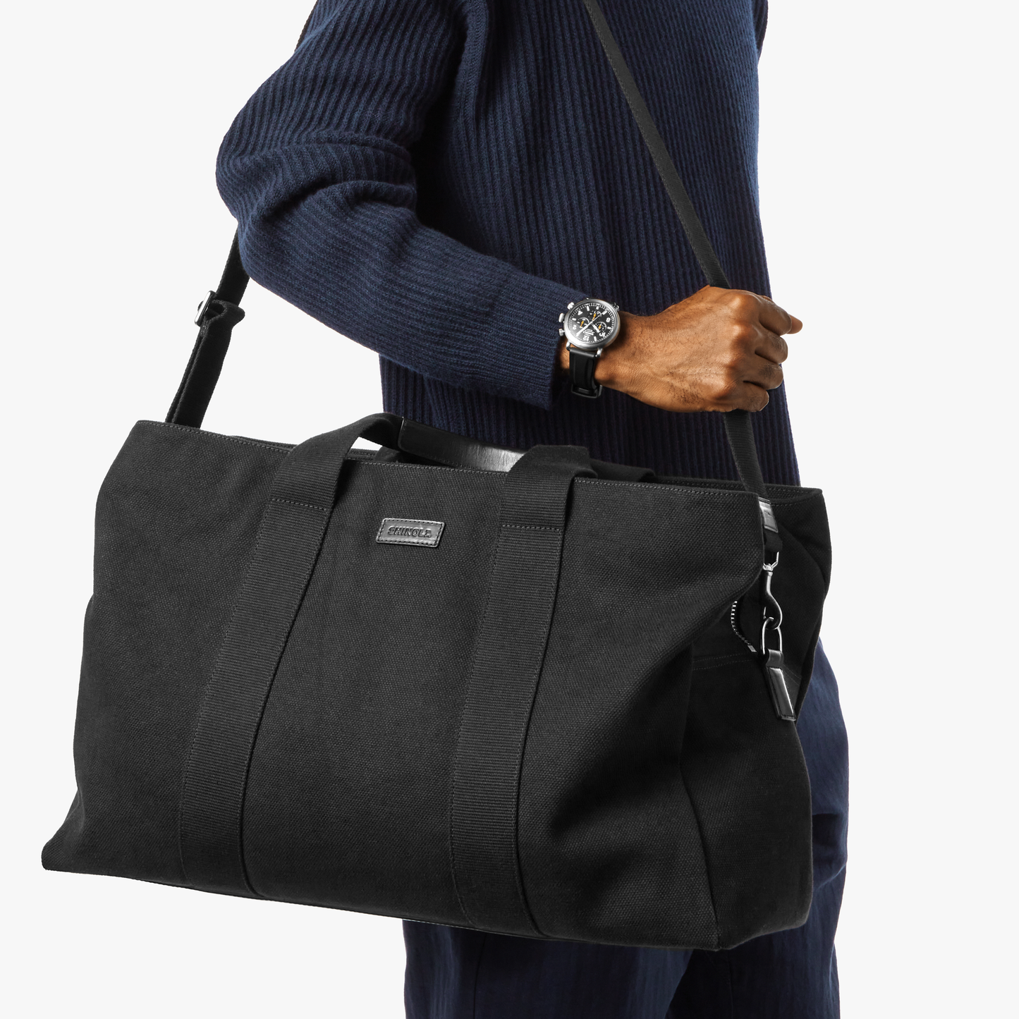 A person in a navy sweater and watch carries the Shinola Runwell Canvas Duffle Bag - Black, featuring hand and shoulder straps—ideal as a TSA carry-on. The background is white.