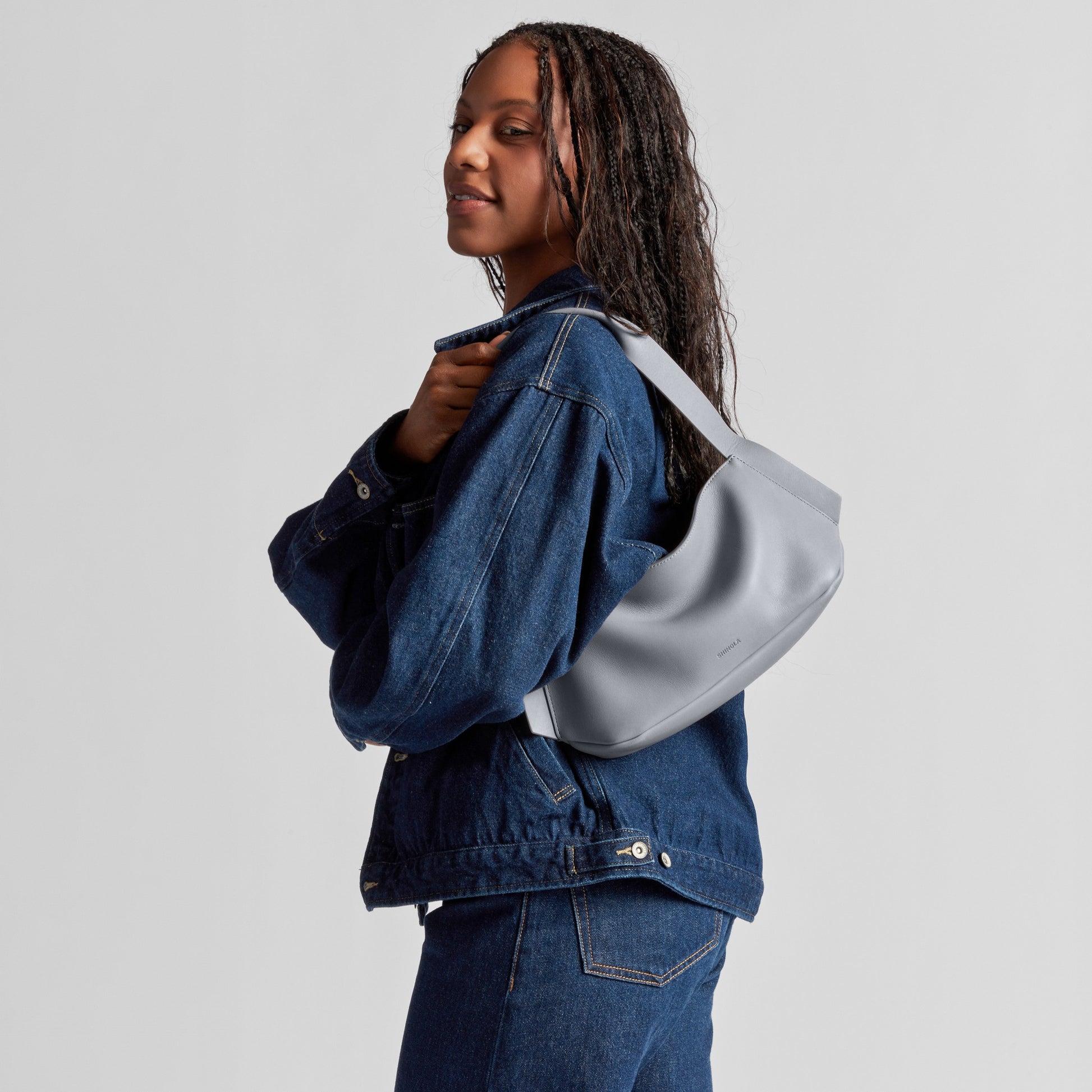 A woman in a denim jacket and jeans stands sideways, smiling softly, with the Shinola Small Derby Shoulder Bag in Mist slung over her shoulder against a plain light background.