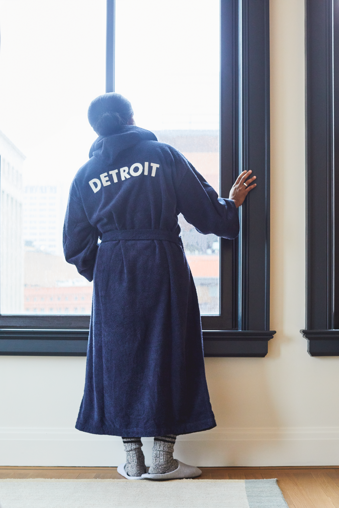 A person in a Shinola Hotel Detroit Robe - Navy by Shinola stands indoors, hand on the window frame, gazing out at city buildings through a large window.