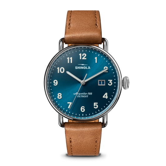 The Shinola Canfield Watch features an astro blue dial with white numerals, a date display, a stainless steel case, and a tan leather strap, shown against a white background.