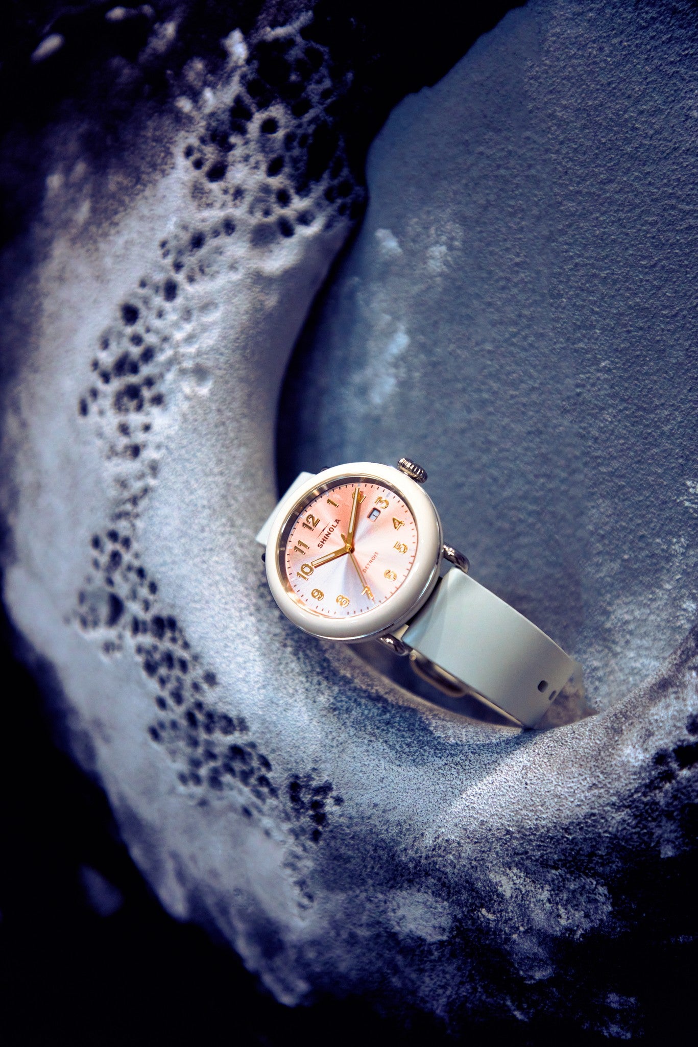 The Shinola Detrola Watch - Nude, showcasing classic American design with a nude band and rose gold face, is displayed on a textured gray surface dusted with white powder.