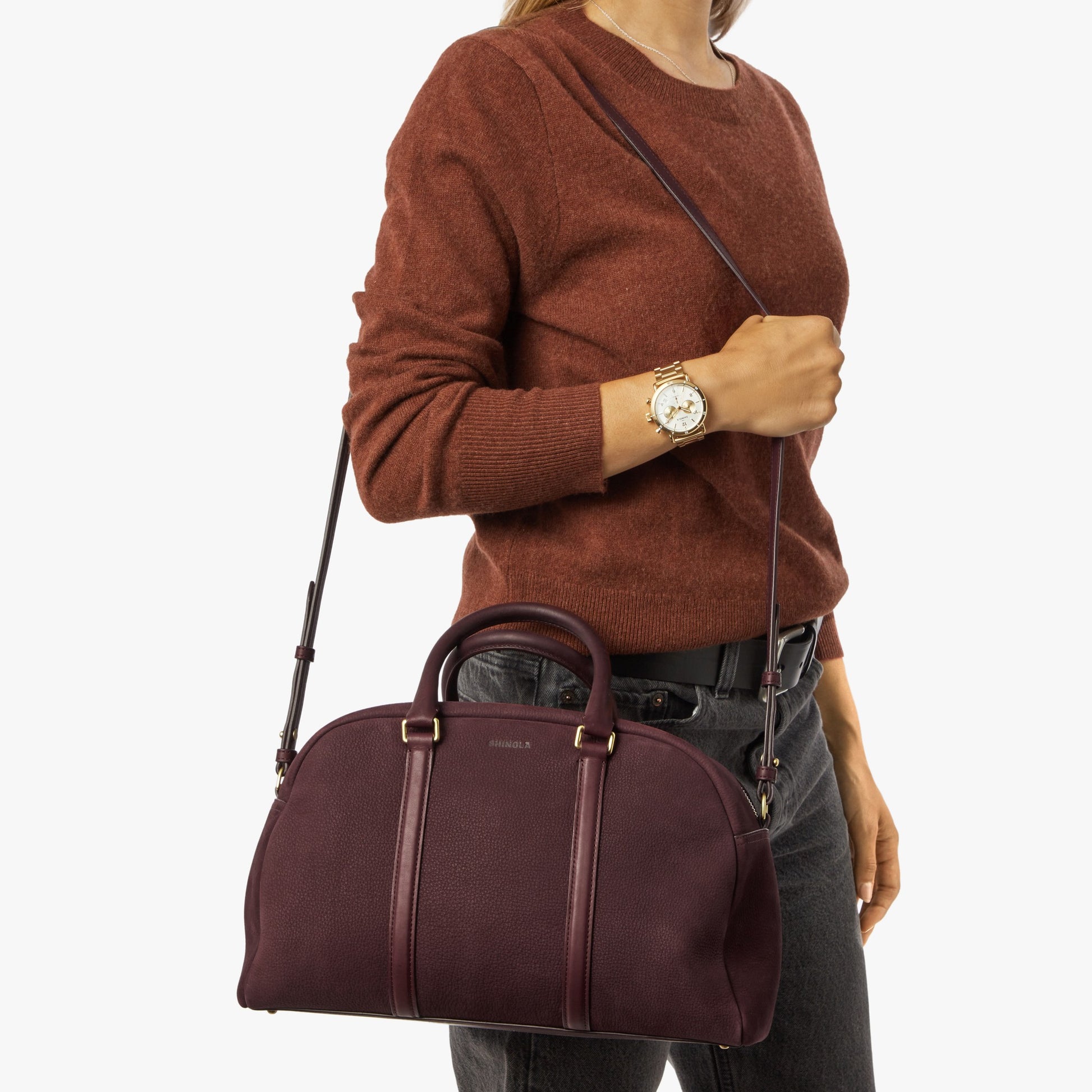 A woman in a rust sweater and dark jeans holds the Shinola Runwell Satchel - Merlot, a women's crossbody bag with shoulder strap and handles. She wears a watch on her left wrist. The background is plain white.