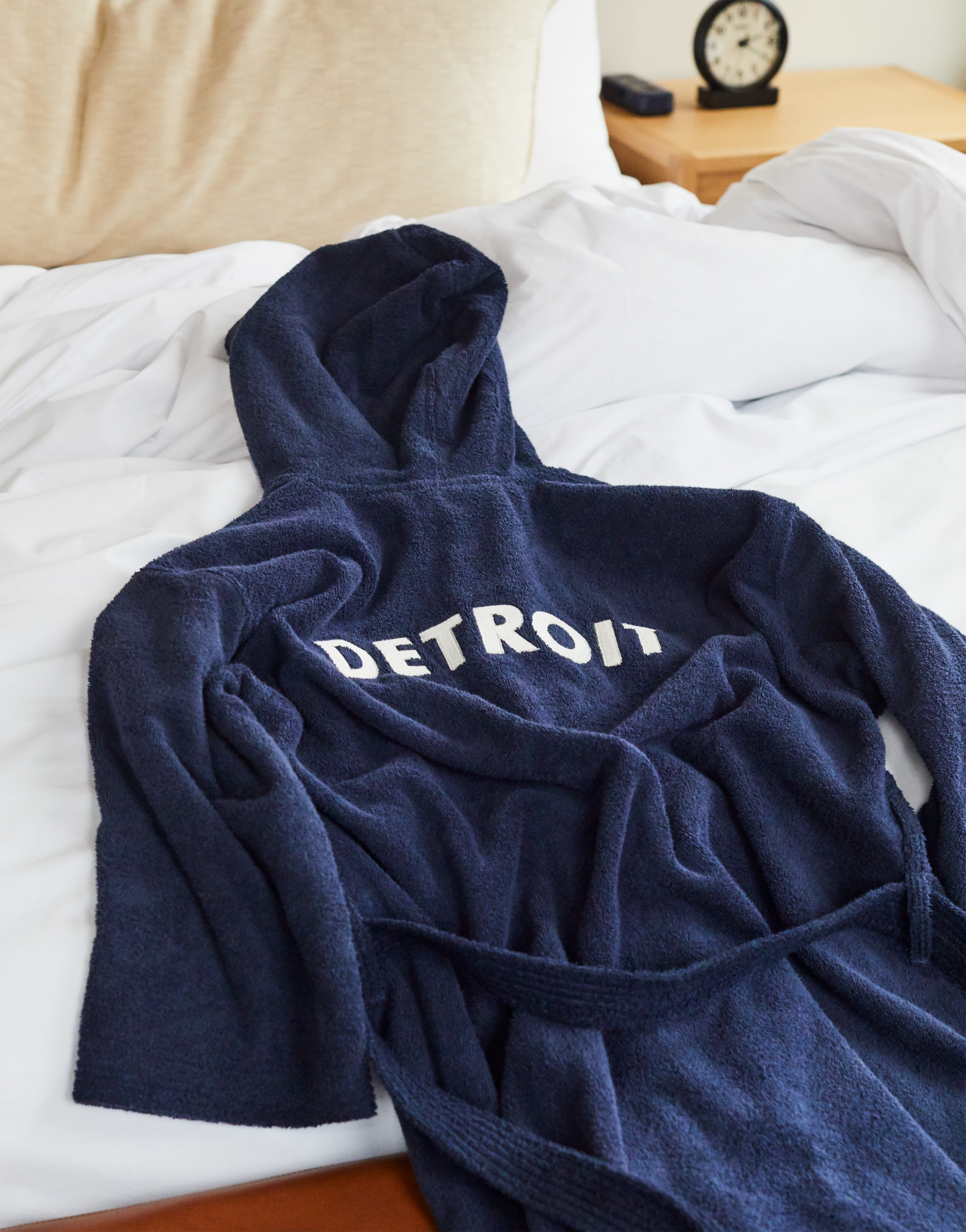 A Shinola Hotel Detroit Robe - Navy by Shinola, crafted from ultra-plush cotton, is neatly displayed on a made white bed in a cozy bedroom, with a pillow and alarm clock in the background.