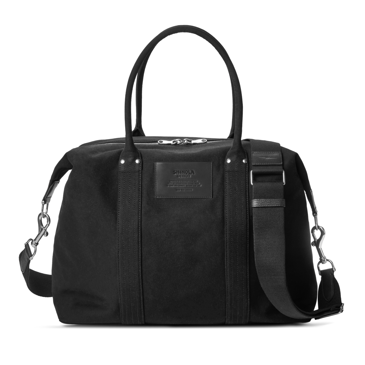 The Shinola Runabout Holdall Canvas Bag in black features cotton canvas, two short handles, a detachable crossbody strap, a zippered top, a subtle front logo patch, and silver hardware accents.