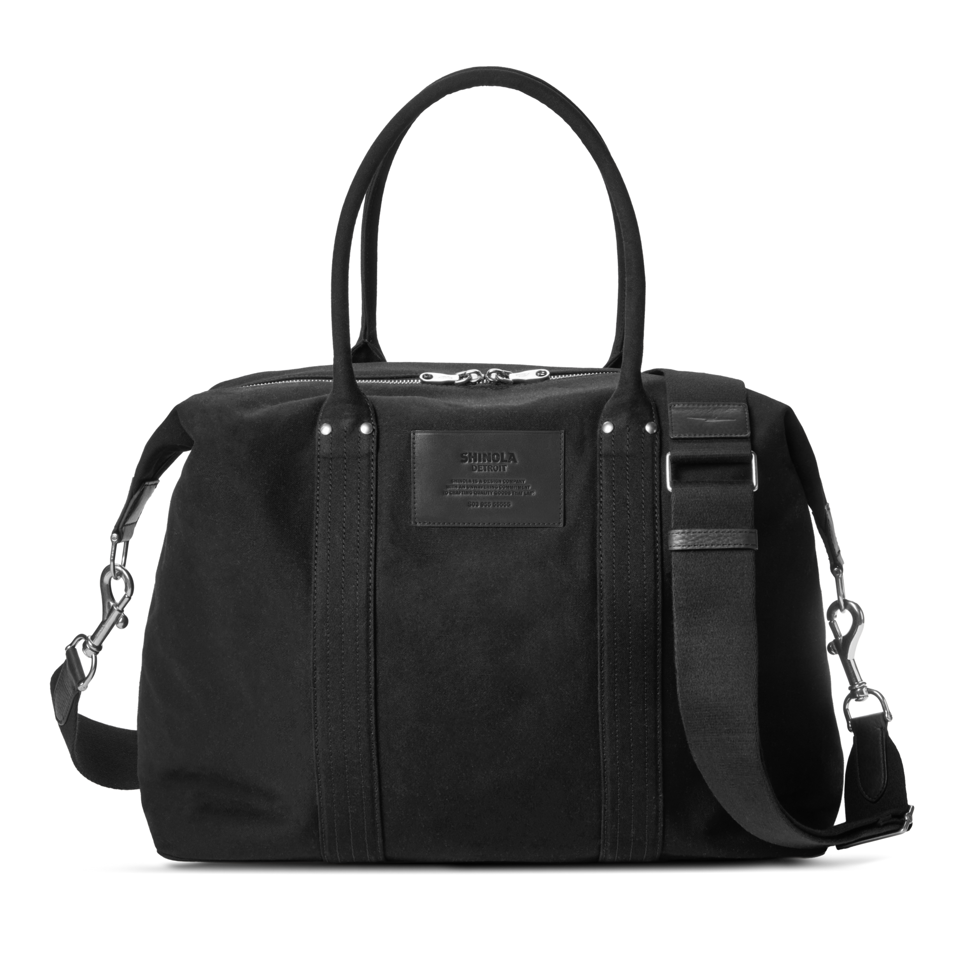 The Shinola Runabout Holdall Canvas Bag in black features cotton canvas, two short handles, a detachable crossbody strap, a zippered top, a subtle front logo patch, and silver hardware accents.