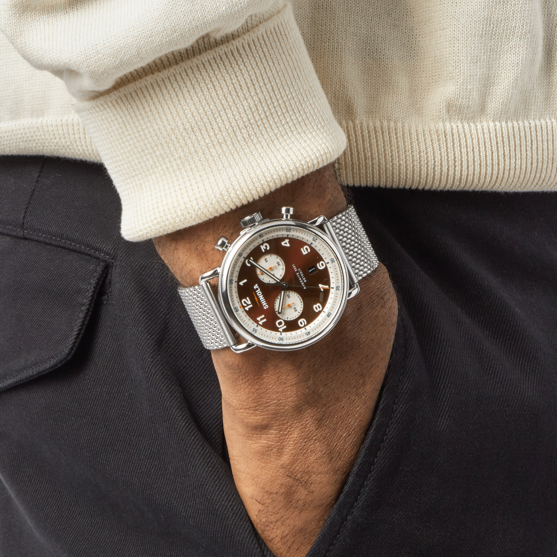 A person in a cream sweater and black pants has their left hand in a pocket, showing off a Shinola Canfield C56 Chronograph Watch - Whiskey with a brown face, two subdials, and a stainless steel mesh bracelet.