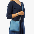 A woman in a navy sweater and blue skirt carries the Shinola Mini Pocket Crossbody Bag - River, featuring a flap and silver hardware. She also wears a silver wristwatch against a plain white background.