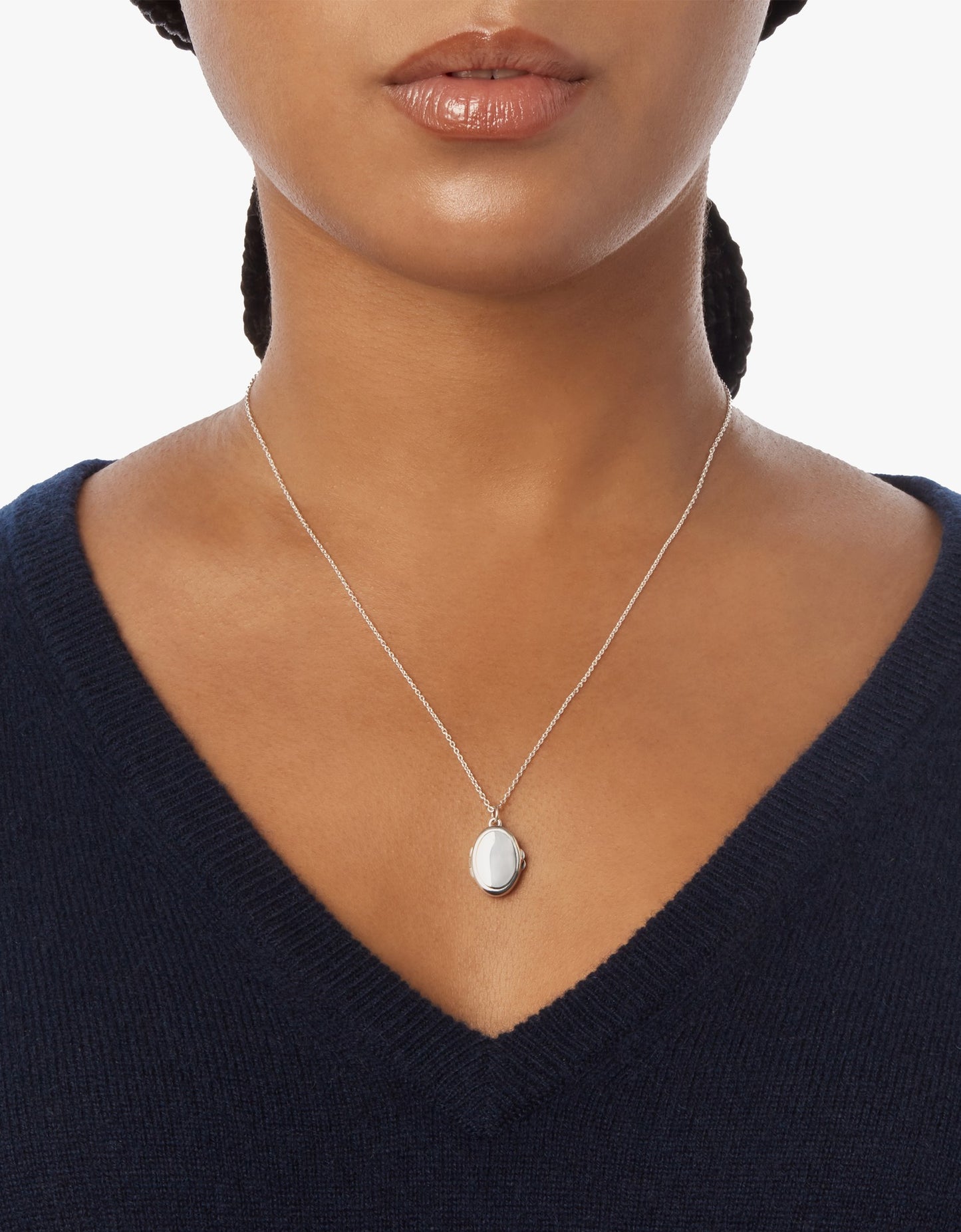 A woman wears a navy V-neck sweater and the Shinola Heirloom Petite Locket Necklace in sterling silver, featuring an oval pendant, shown from her shoulders to just above her lips.