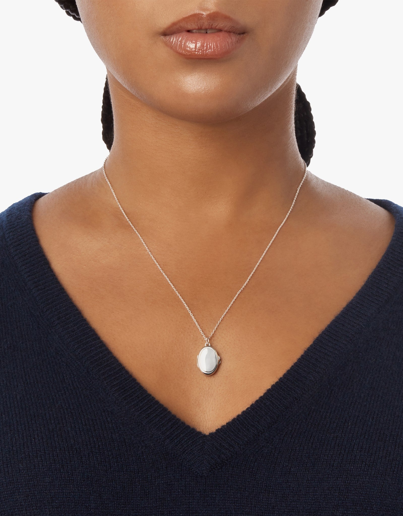 A woman wears a navy V-neck sweater and the Shinola Heirloom Petite Locket Necklace in sterling silver, featuring an oval pendant, shown from her shoulders to just above her lips.