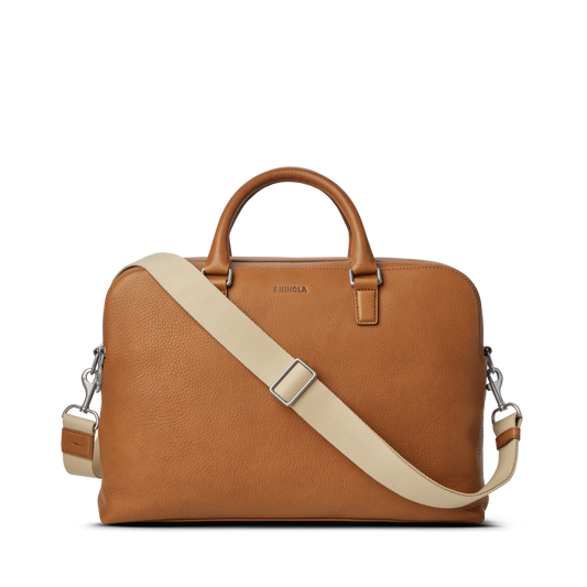 The Shinola Double Zip Briefcase - Tan, crafted from natural grain leather, features short handles, a detachable beige shoulder strap, an extra compartment, silver hardware, and a simple elegant design against a white background.