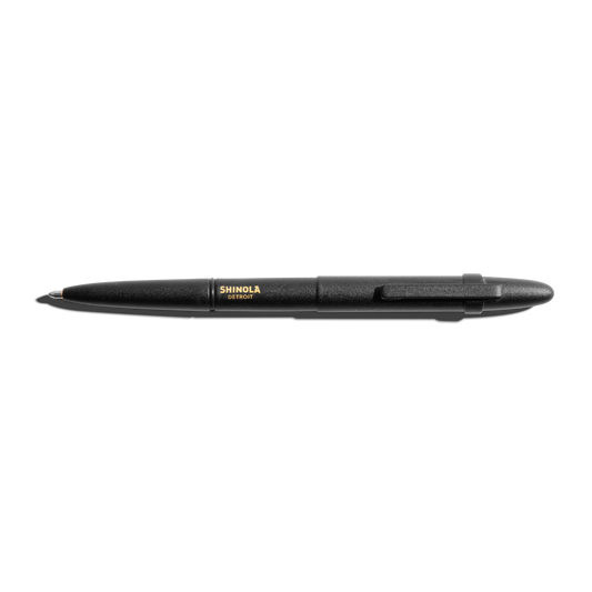The Shinola + Fisher Bullet Space Pen - Matte Black, with gold SHINOLA DETROIT branding and a clip, is shown against a white background—designed to write underwater and in extreme conditions.