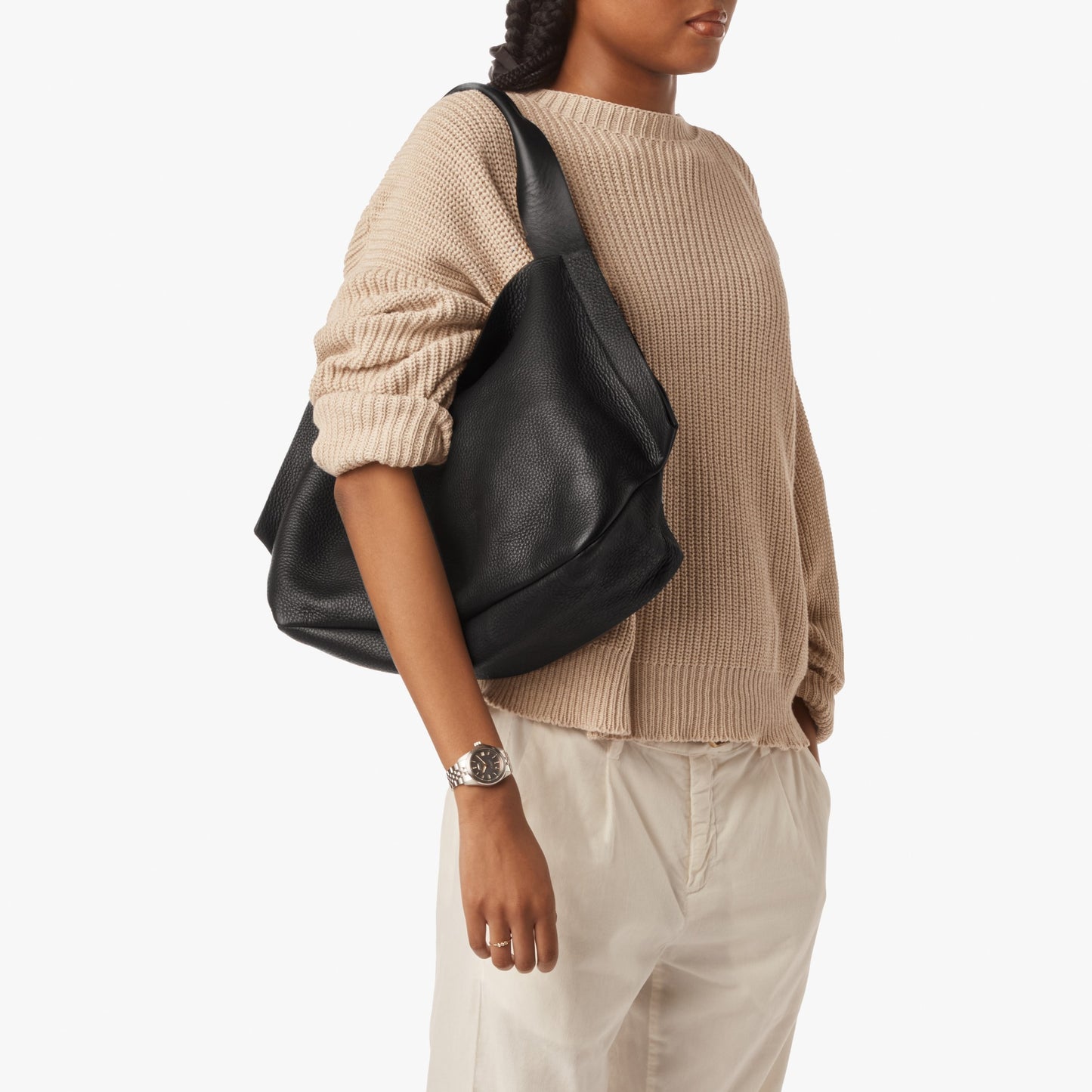 A person wearing a beige knit sweater and light pants carries the Shinola Derby Shoulder Bag - Black, which features a large open compartment and leather card pocket. They also wear a silver wristwatch. The background is plain white.