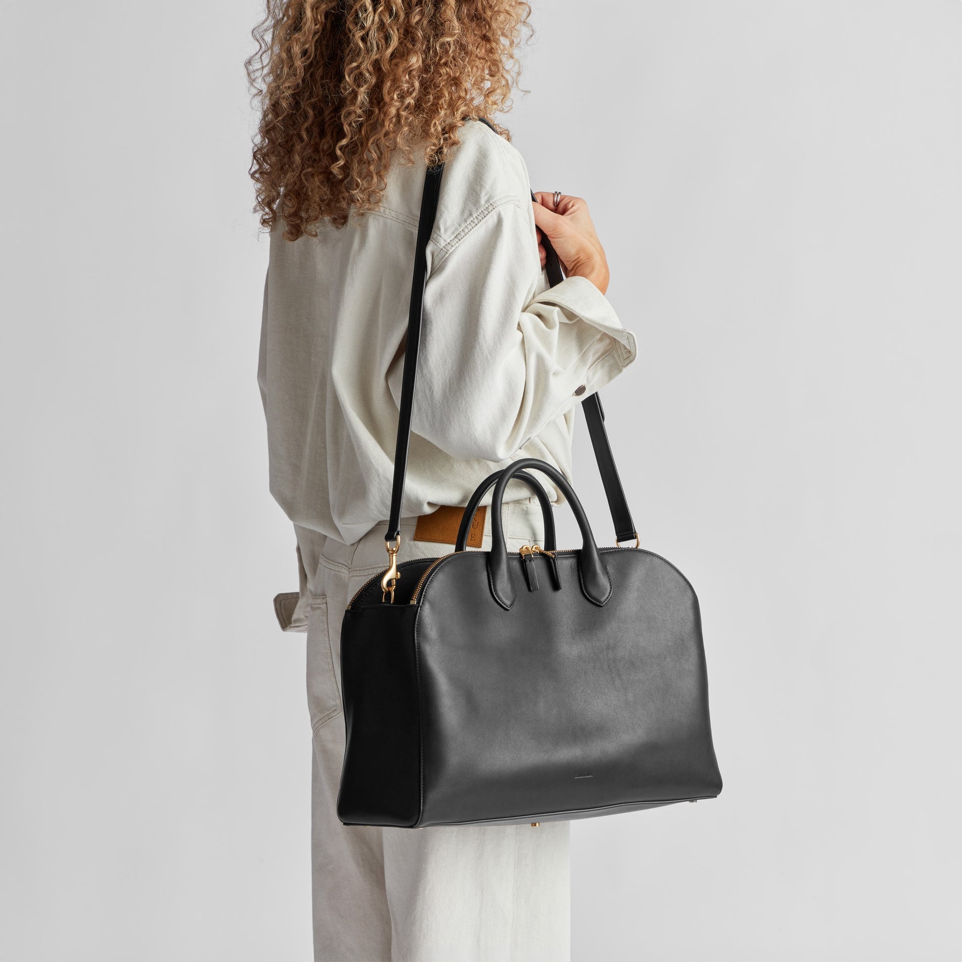 A person with curly hair, in light-colored, loose clothing, carries a large black Shinola Bixby Work Satchel with a shoulder strap against a plain light background.