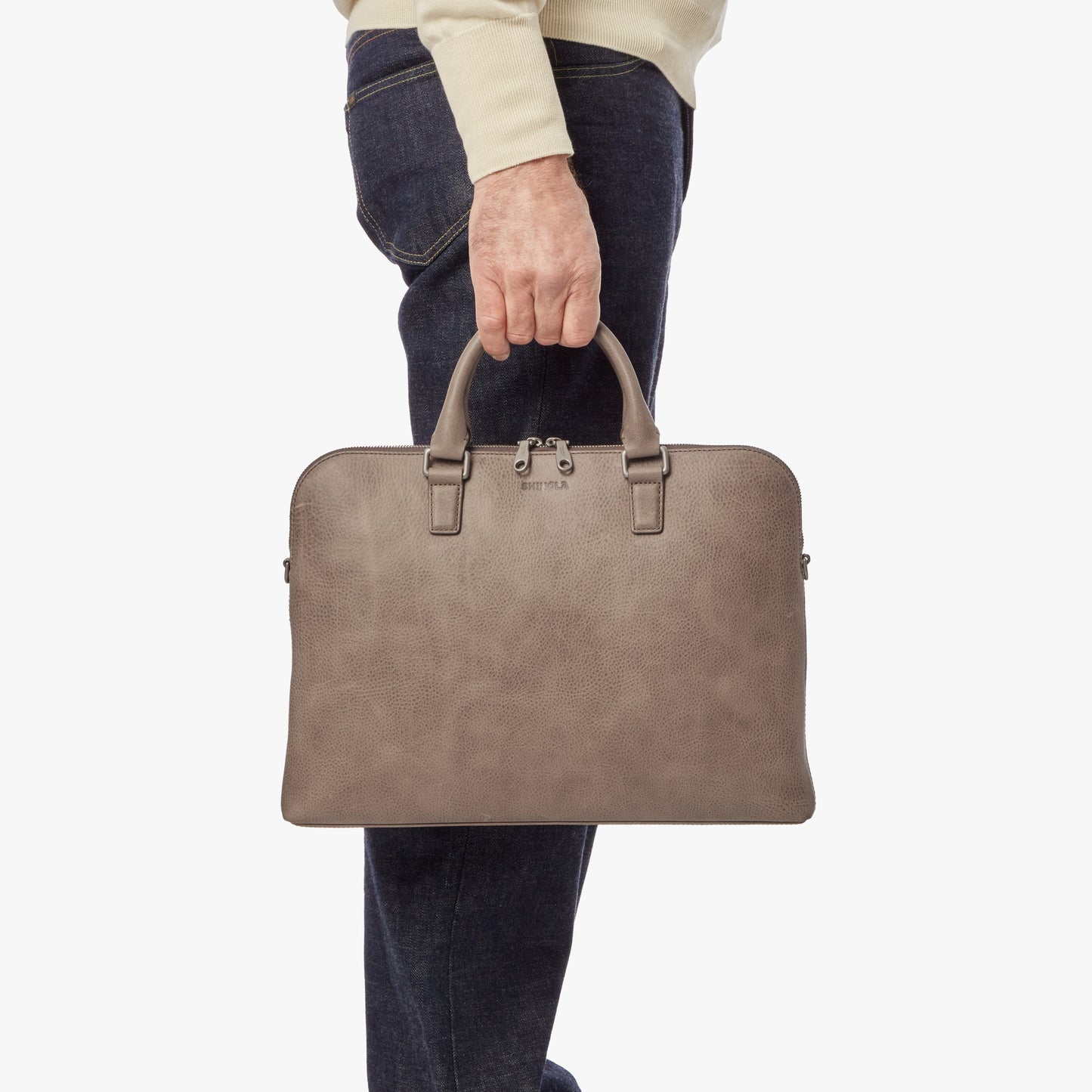 A person in dark jeans and a light sweater holds a stone leather Portfolio Briefcase by its handles, with the Shinola brand name visible on the front, against a plain white background.