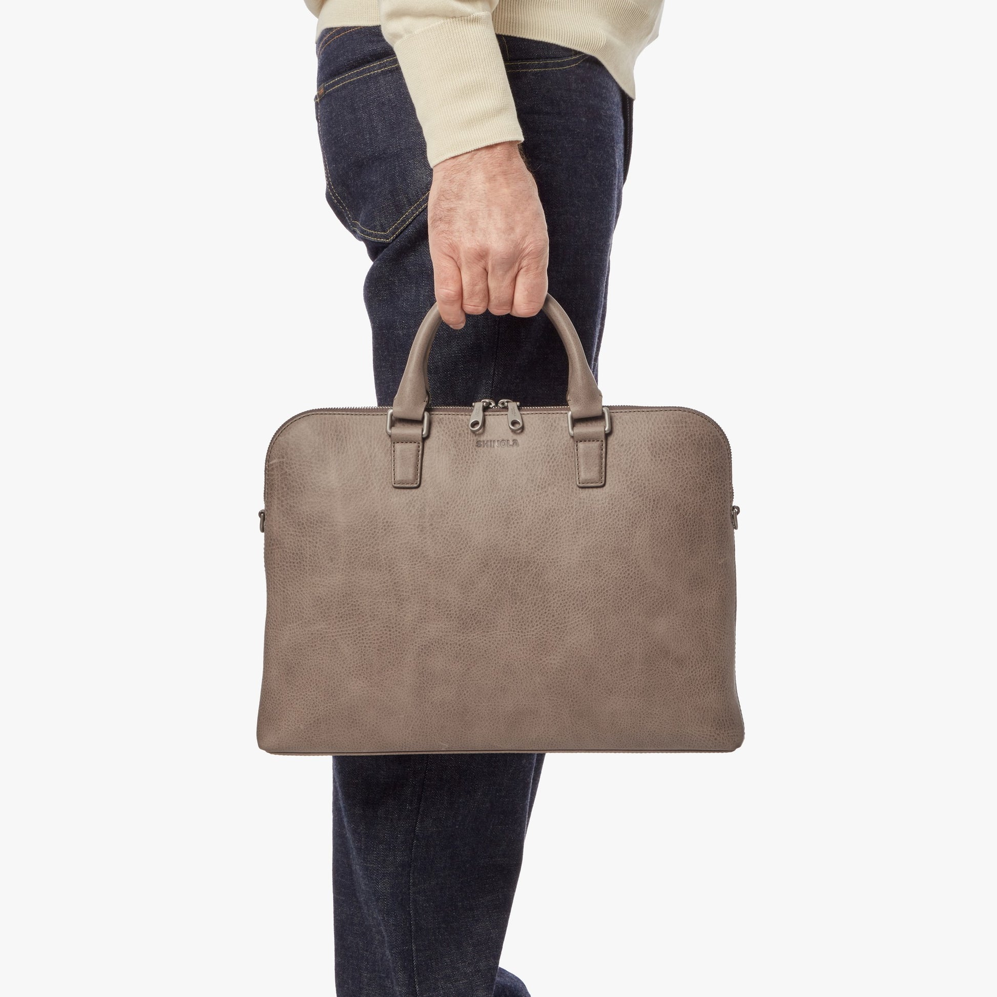 A person in dark jeans and a light sweater holds a stone leather Portfolio Briefcase by its handles, with the Shinola brand name visible on the front, against a plain white background.