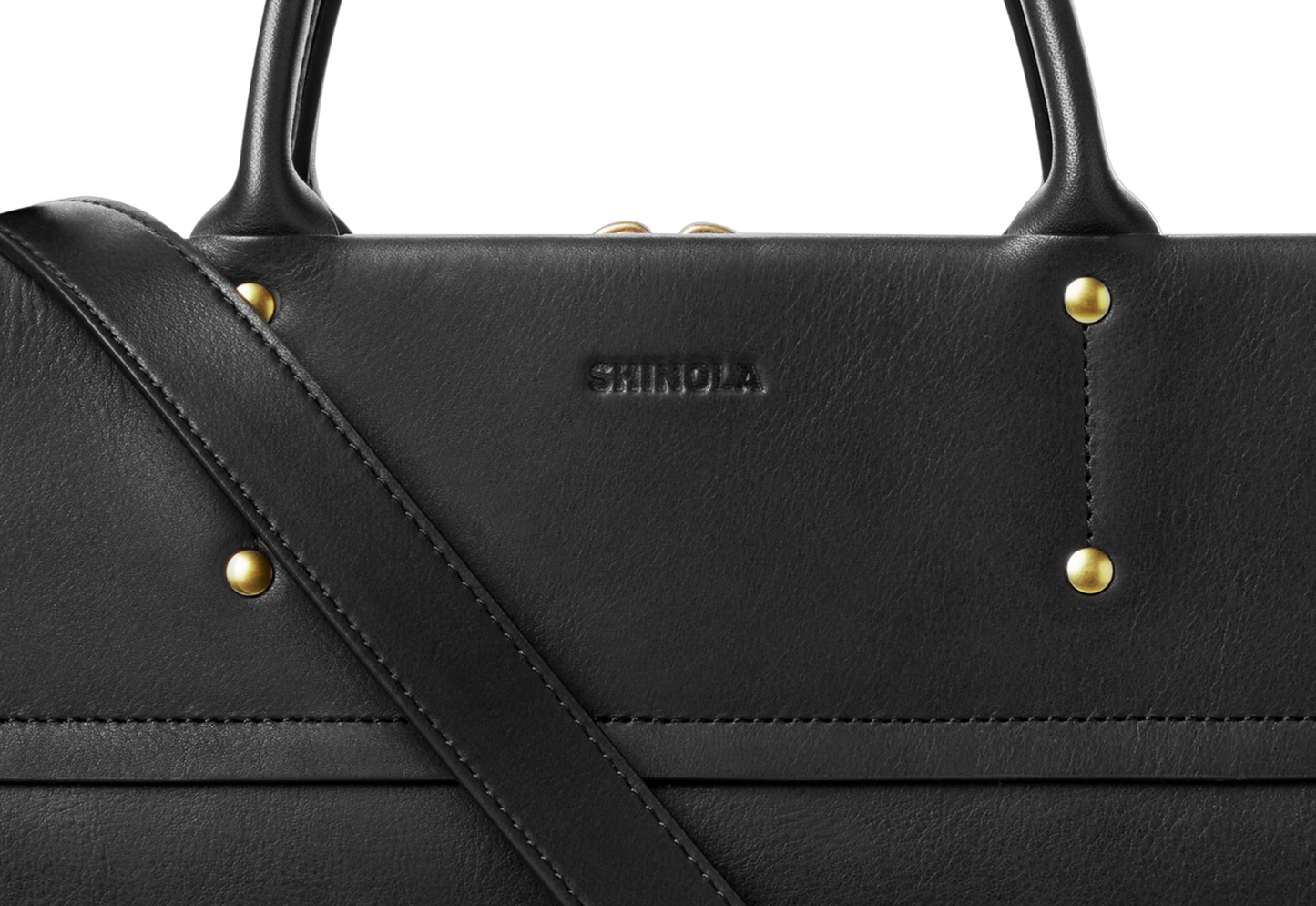Close-up of the Shinola Day Satchel - Black, a versatile leather bag with gold rivets and “Shinola” embossed on it. Features include sturdy top handles and a diagonal strap, ideal for on-the-go use against a plain background.