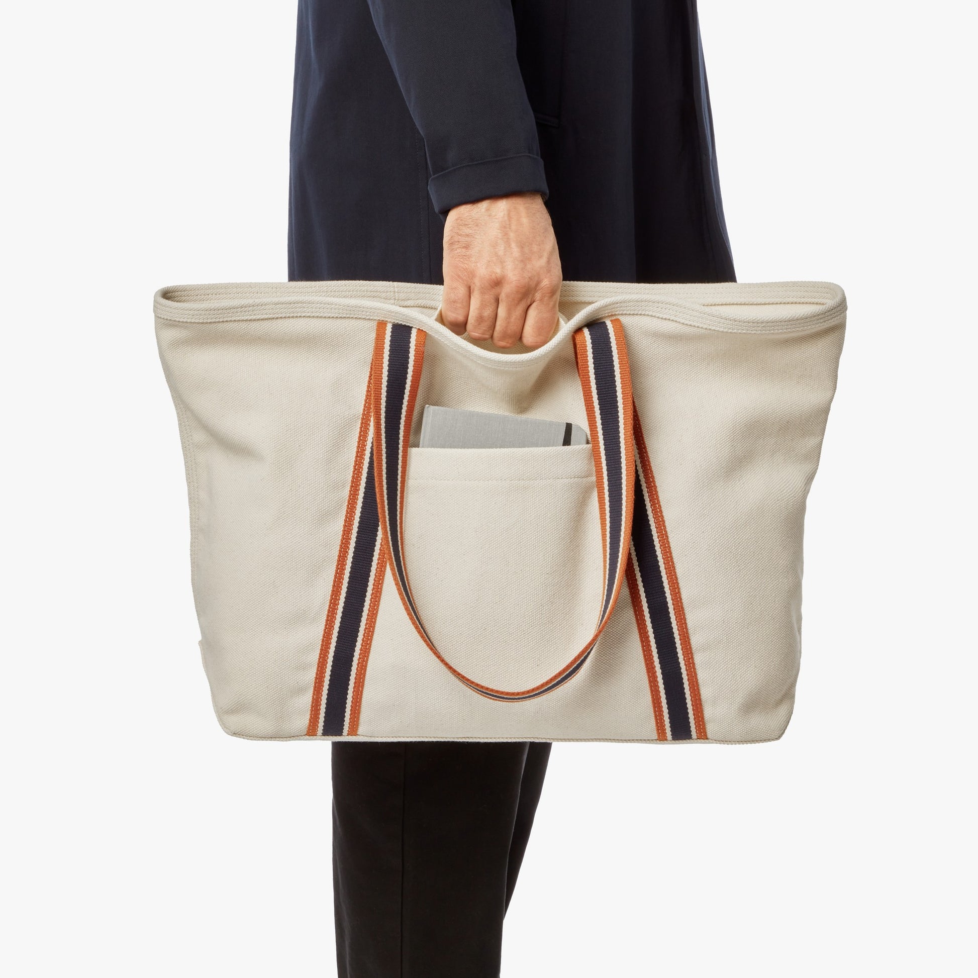 A person in dark clothing holds the Shinola Large Easy Canvas Tote - Natural, which has orange and black striped handles, a spacious main compartment, and an exterior slip pocket with a gray notebook.