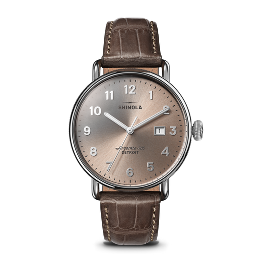 The Shinola Canfield Watch features a 38mm round silver case, tan dial with silver markers, numerals, hands, and date window at 3 o’clock. It comes with a gray-brown American alligator leather strap and runs on the Argonite 705 movement.